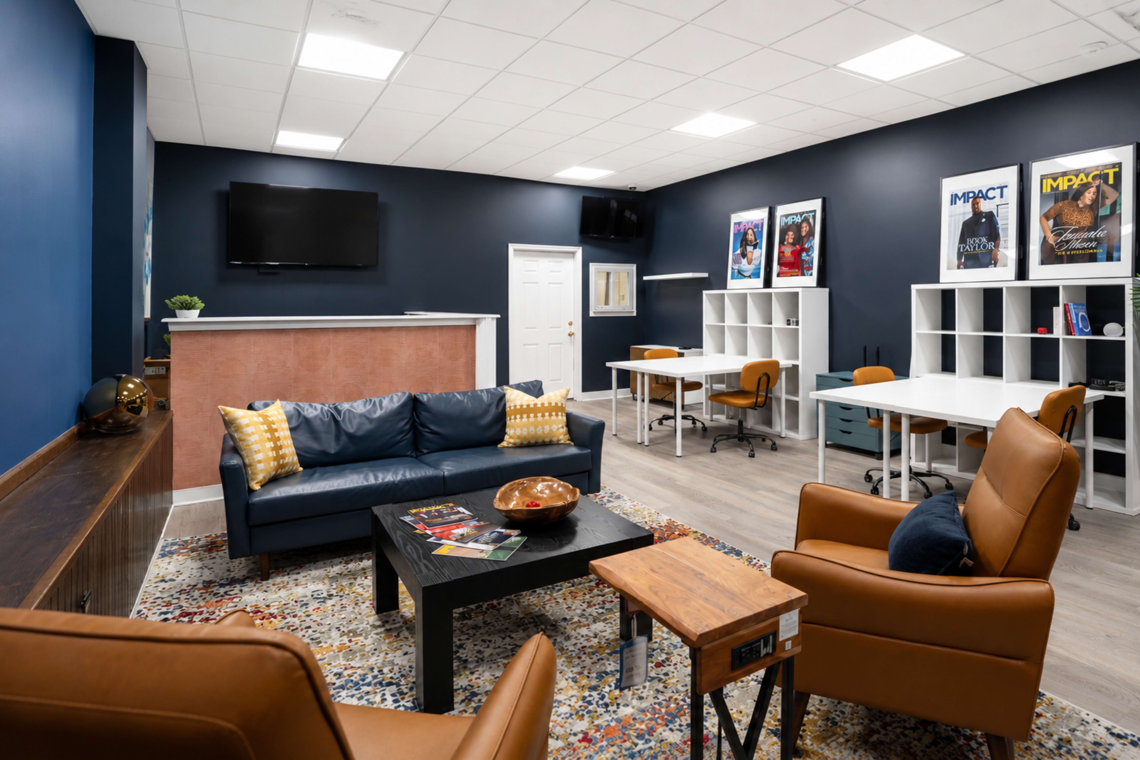 The image shows a modern office lounge area with a blue accent wall, comfortable seating, a coffee table, and workspace stations equipped with desks and chairs.