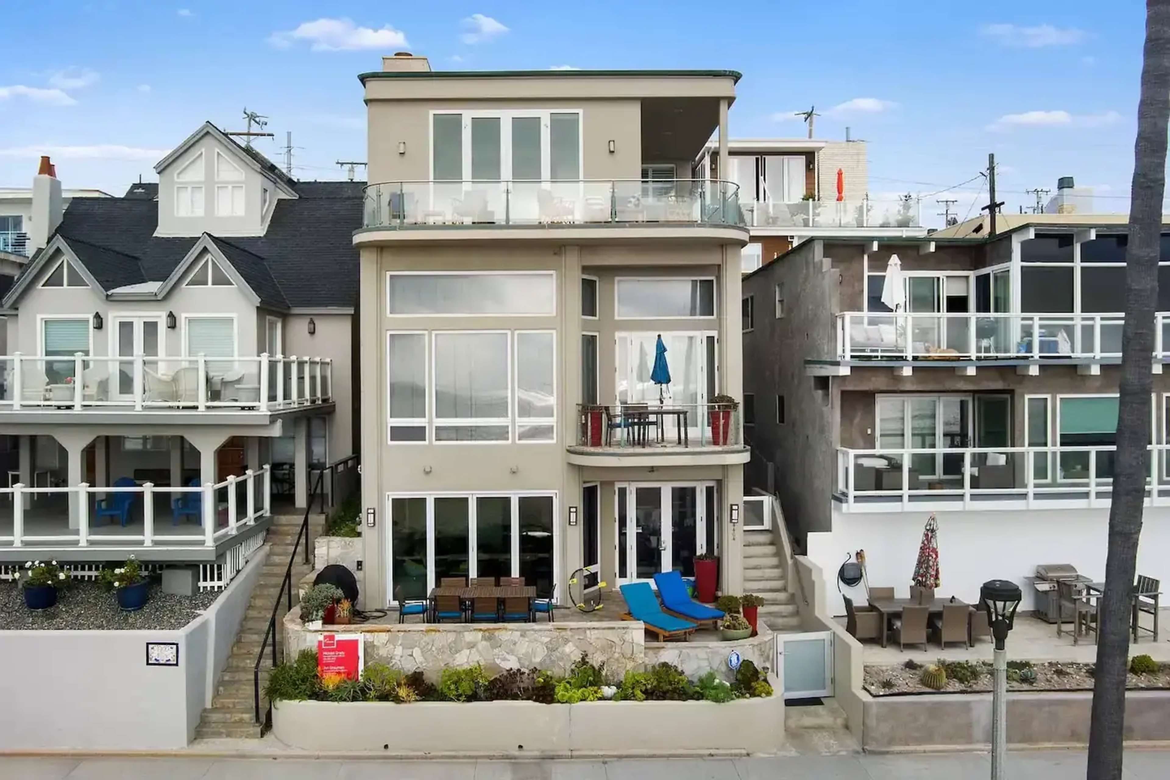 A modern beach house with large glass windows and multiple outdoor seating areas, situated adjacent to other similar homes along a coastal street.