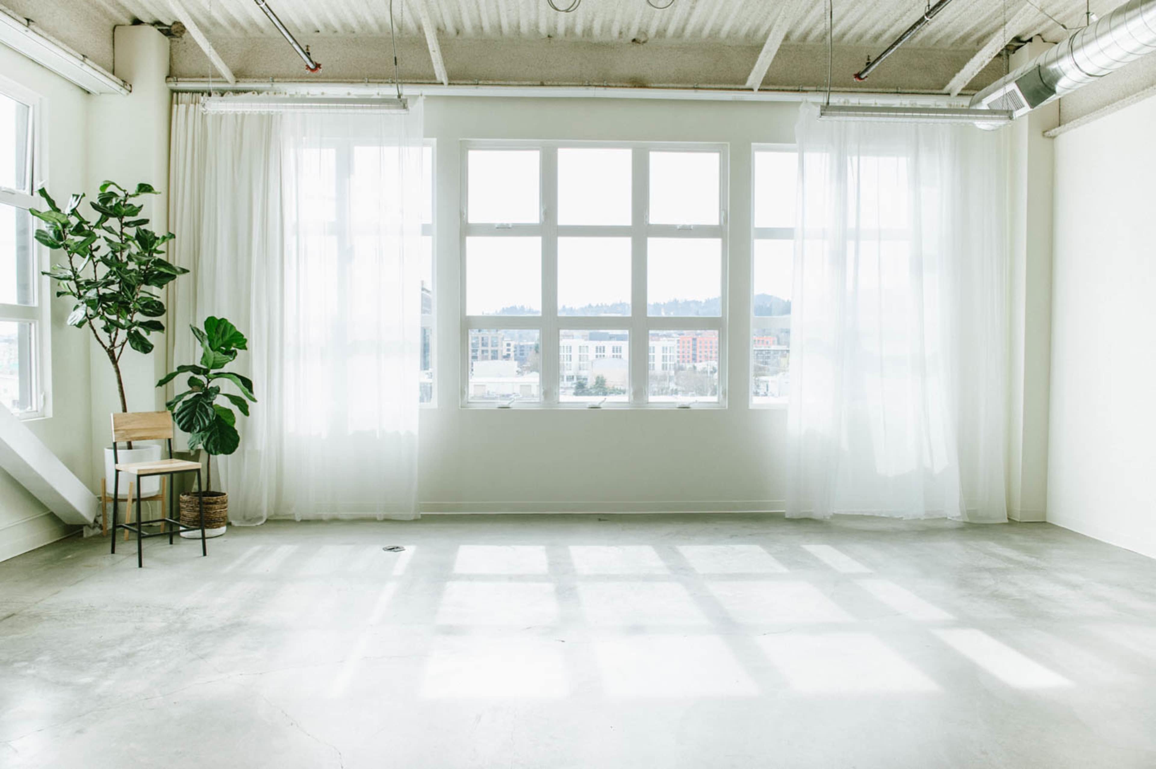 A spacious room features large windows covered with sheer curtains, a potted plant, and a wooden chair against a light-colored wall.