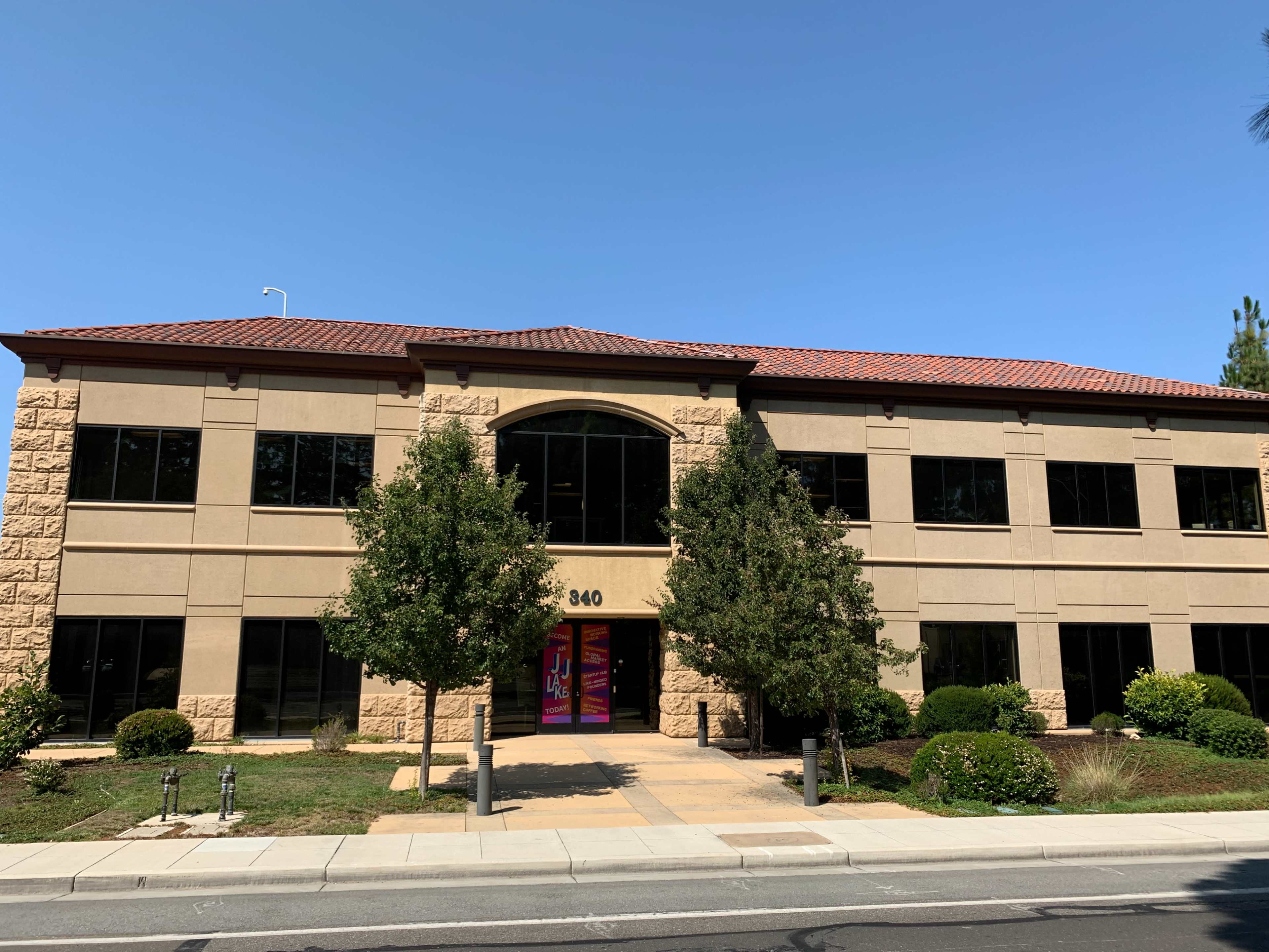 A two-story office building with tan stone exterior and large windows is situated on a landscaped lot.