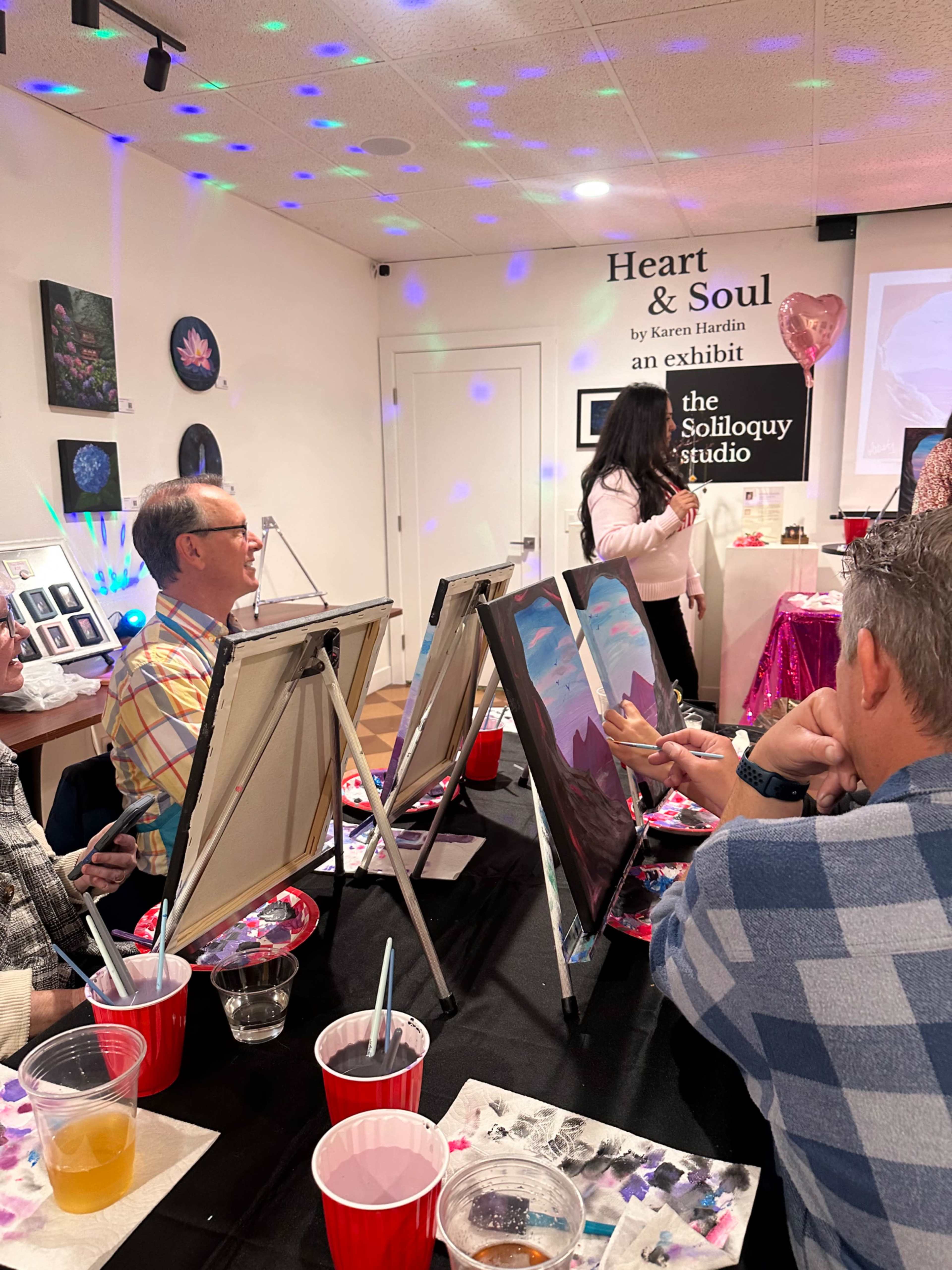 In a studio setting, participants are painting on canvases while an instructor guides them, with colorful lights and decor in the background.
