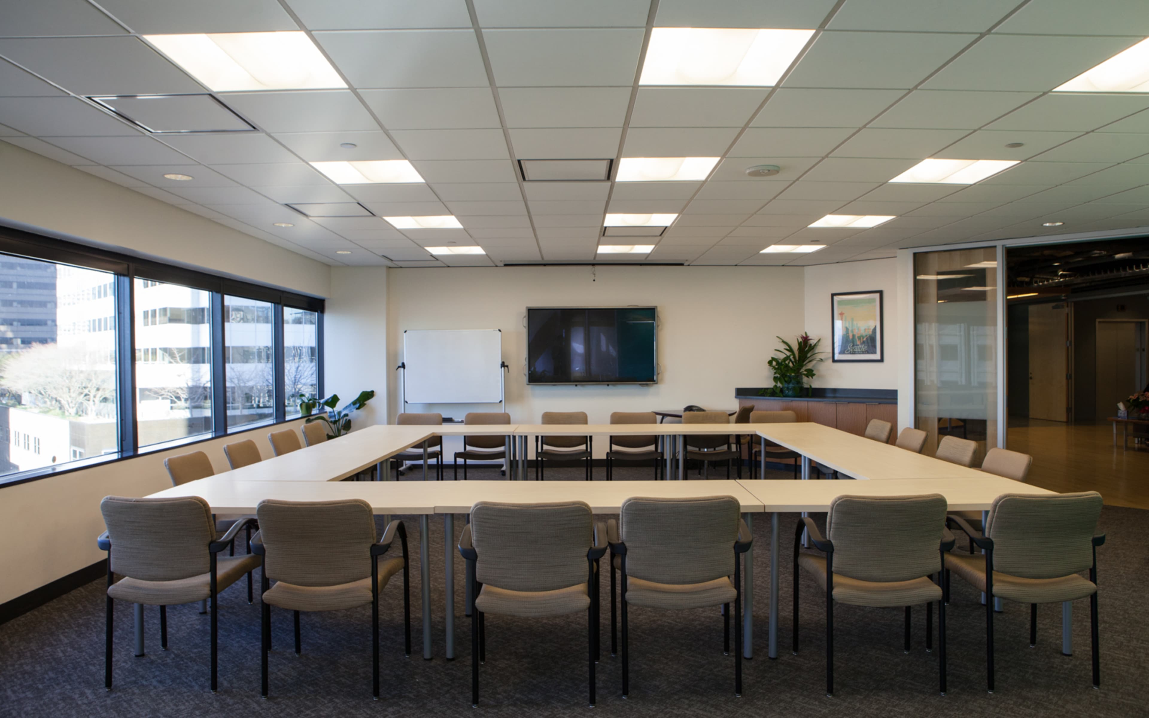 A conference room features a large rectangular table surrounded by chairs, with windows providing natural light and a television mounted on the wall.