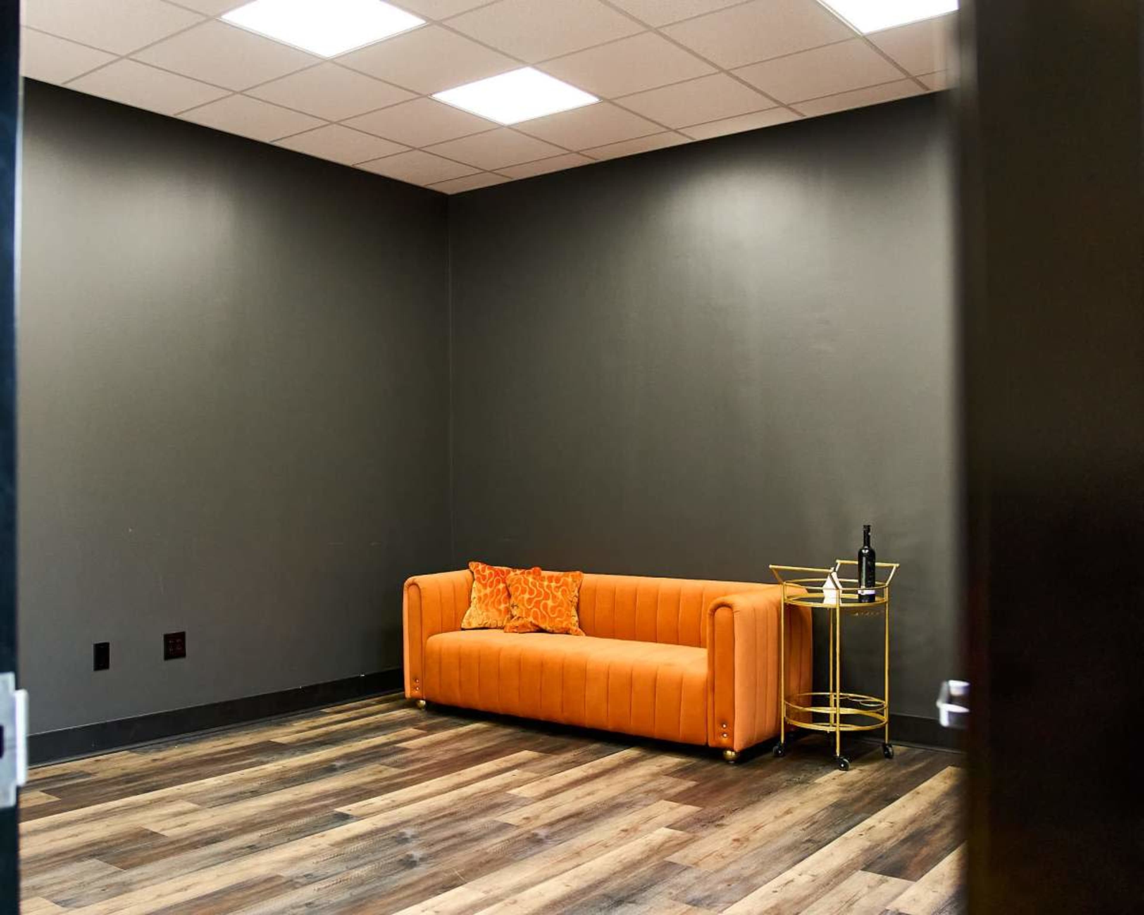 A minimalist room features an orange couch and a small gold cart with beverages against a dark wall.