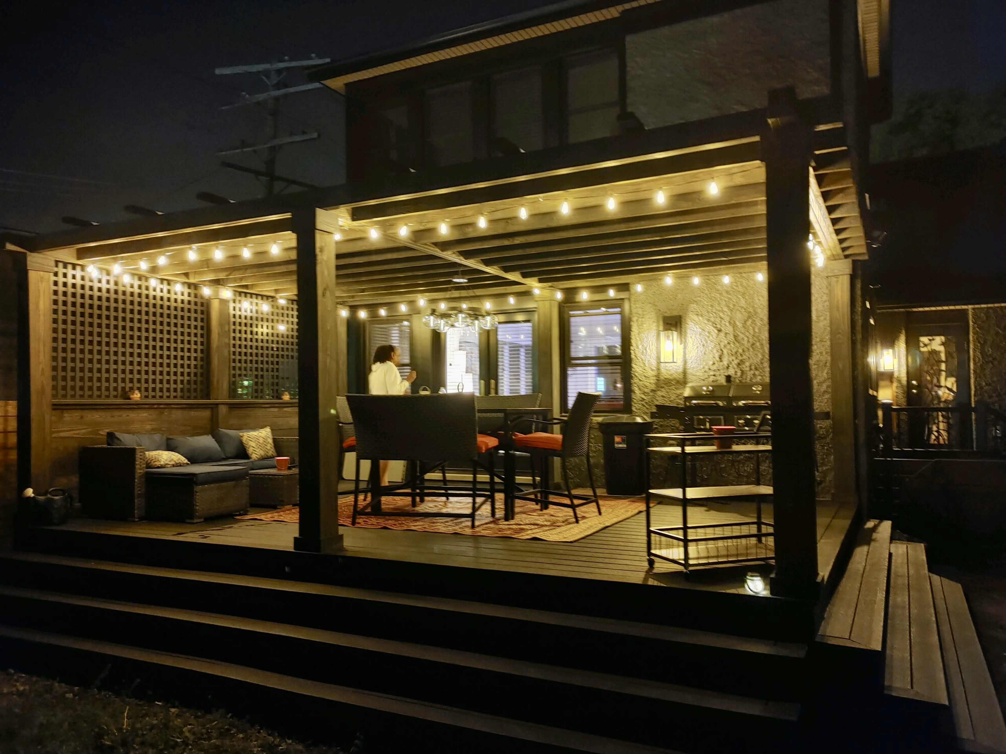 A well-lit outdoor patio features a table and chairs beneath a covered pergola, with string lights illuminating the space at night.