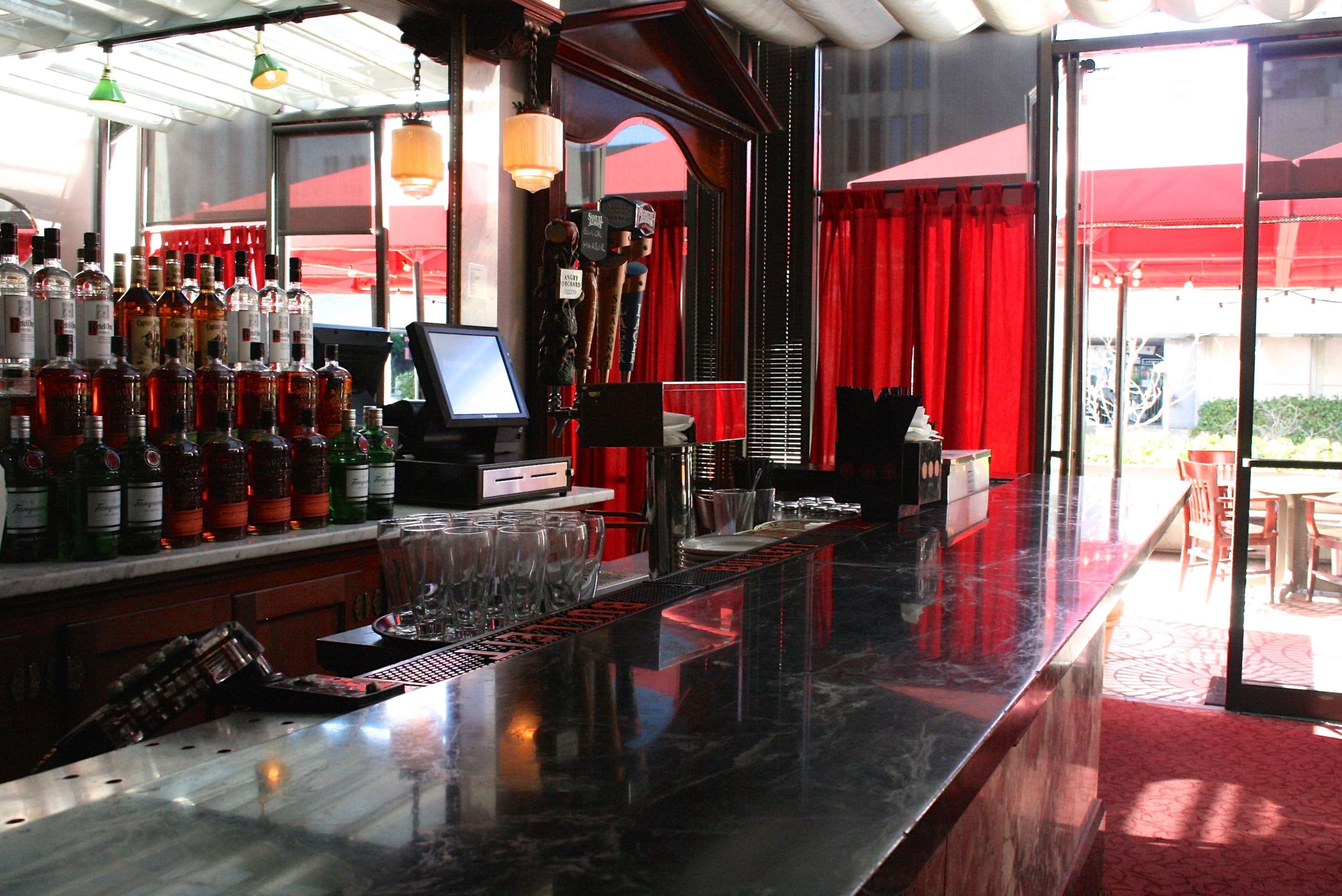A bar with a marble countertop, shelves stocked with various bottles of liquor, and a view of red umbrellas and outdoor seating through glass doors.