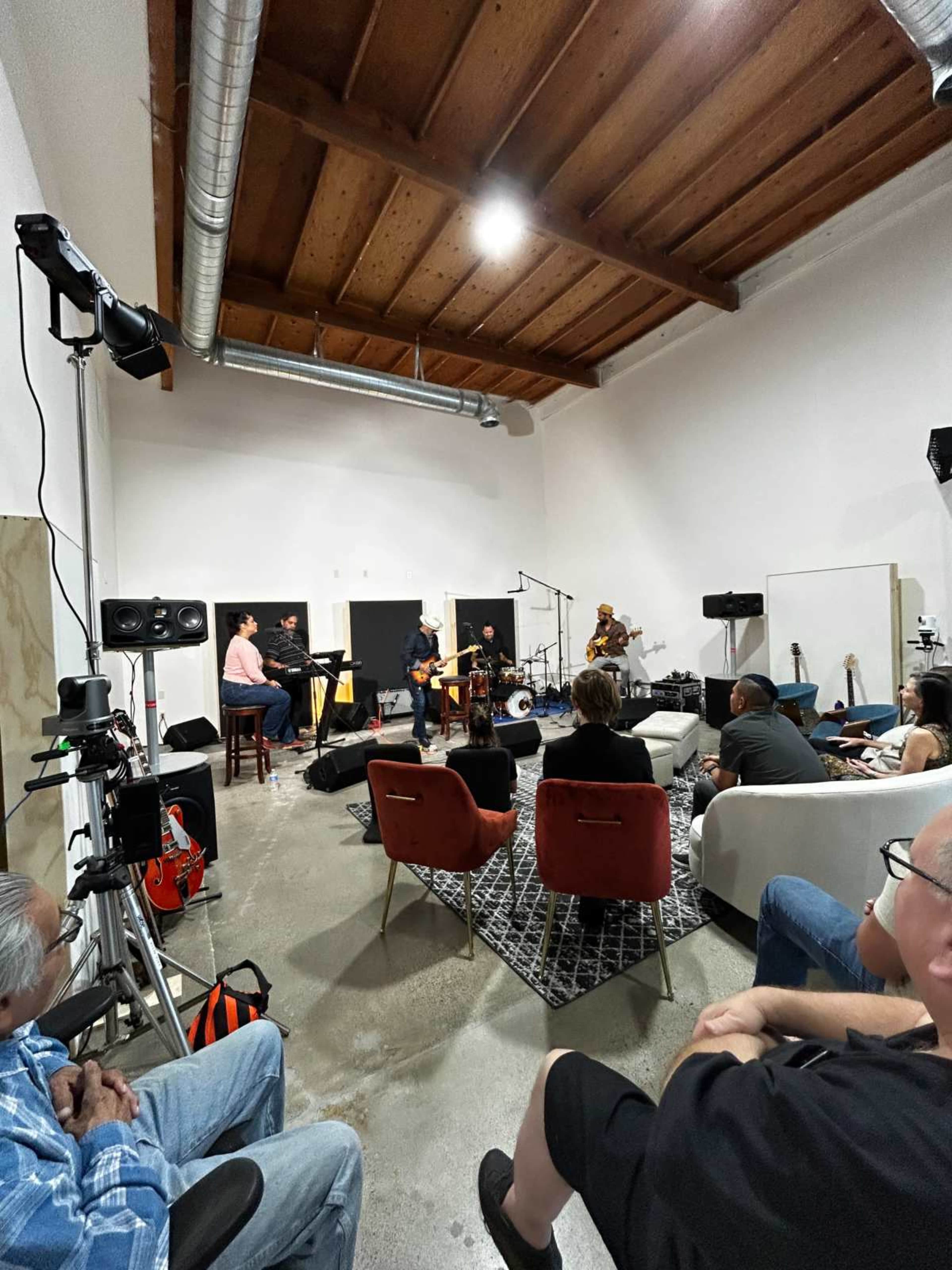 A small group of musicians performs on a stage in a spacious, minimalist room while an audience sits on sofas and chairs.