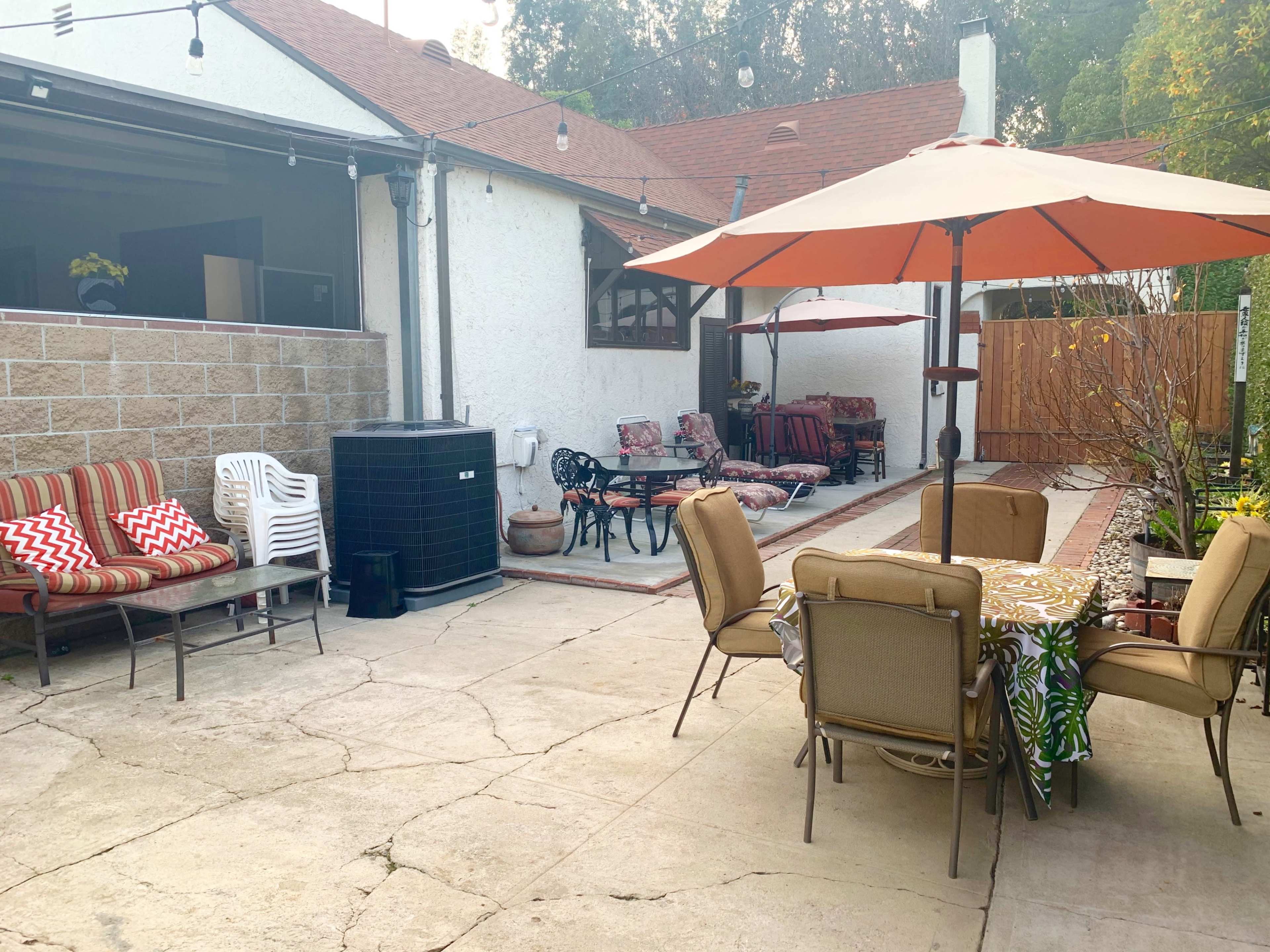 The image shows a patio area with various seating arrangements, including a table and chairs under an umbrella, surrounded by a garden and a wall.