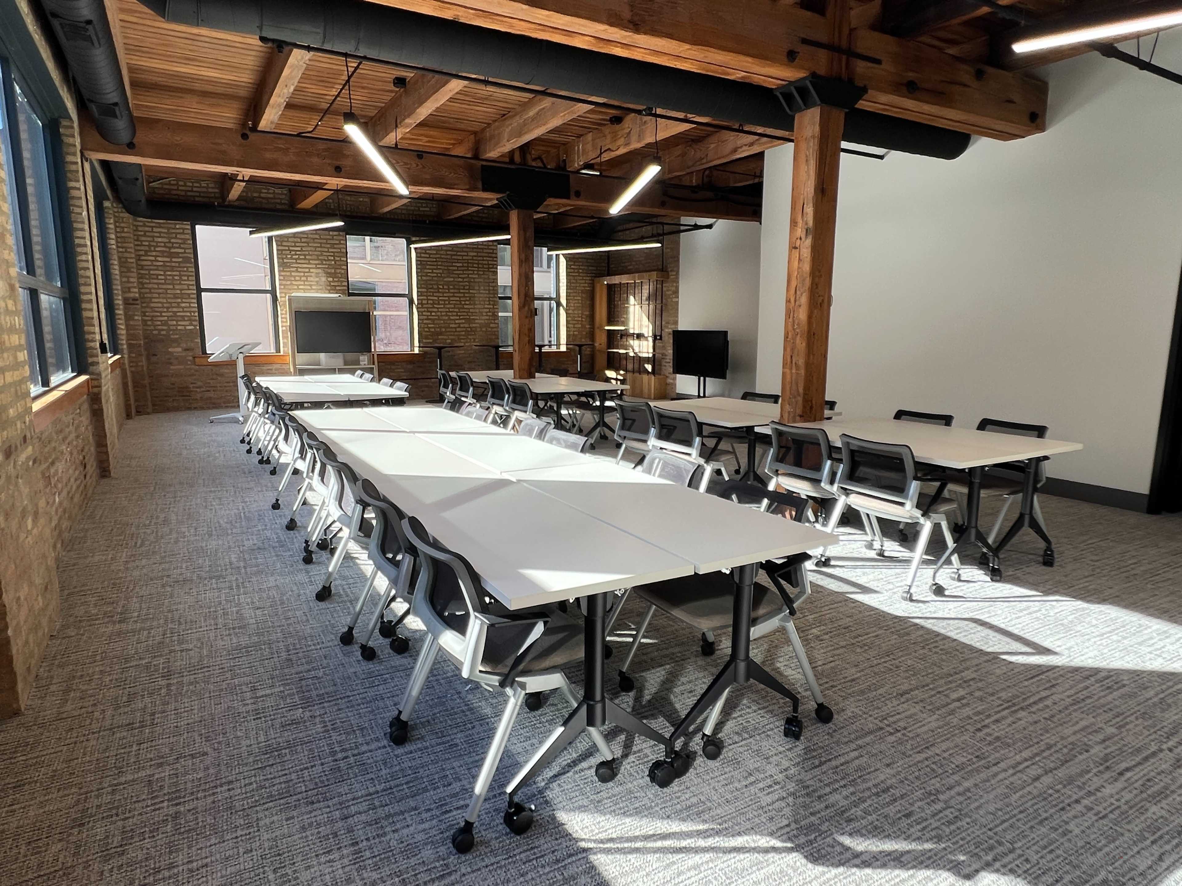 The image shows a modern meeting room with several rows of white tables and rolling chairs arranged in a rectangular layout.