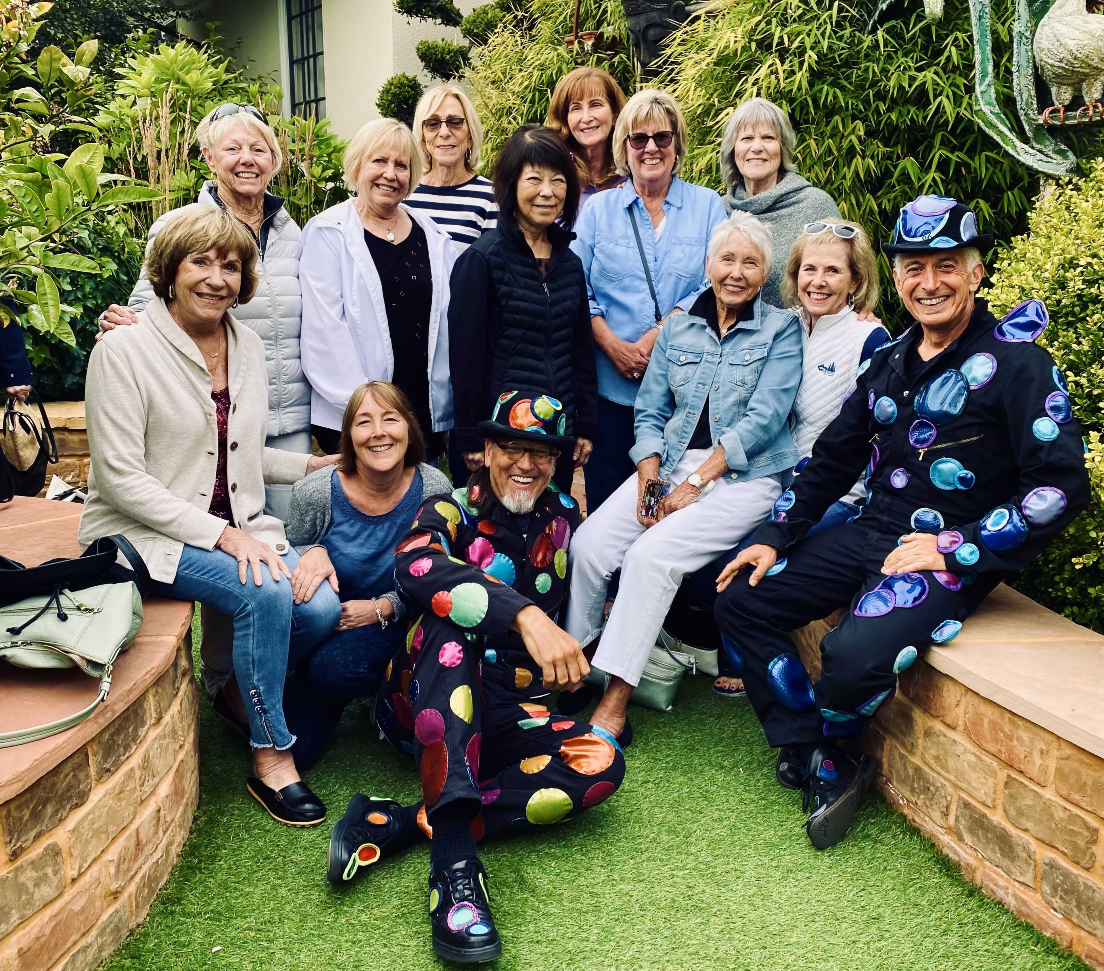 A group of thirteen people poses together in a garden setting, with one individual dressed in a colorful polka-dotted costume.