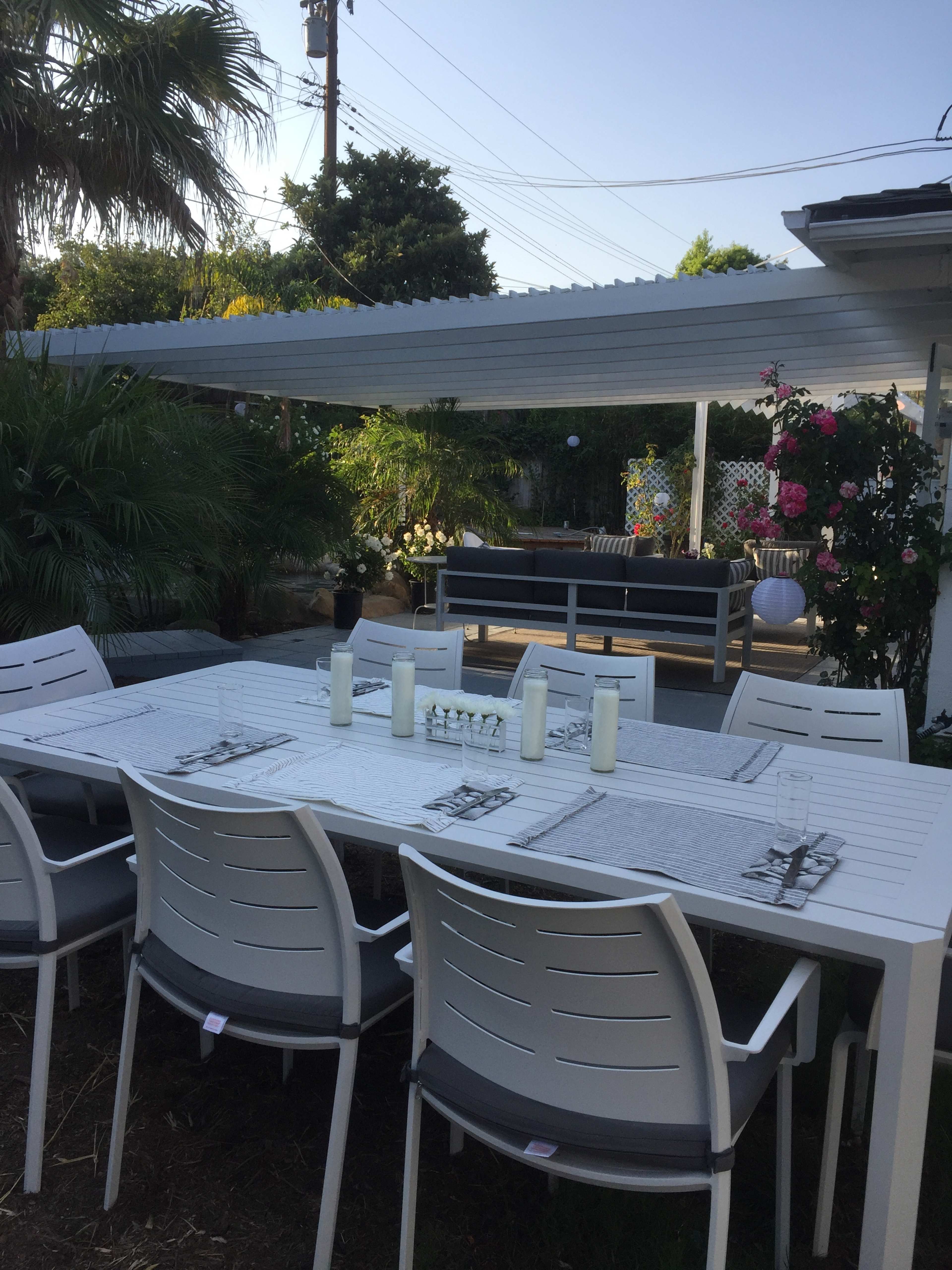 A spacious outdoor dining area with a white table set for six and surrounded by plants, under a pergola and adjacent to a lounge area.