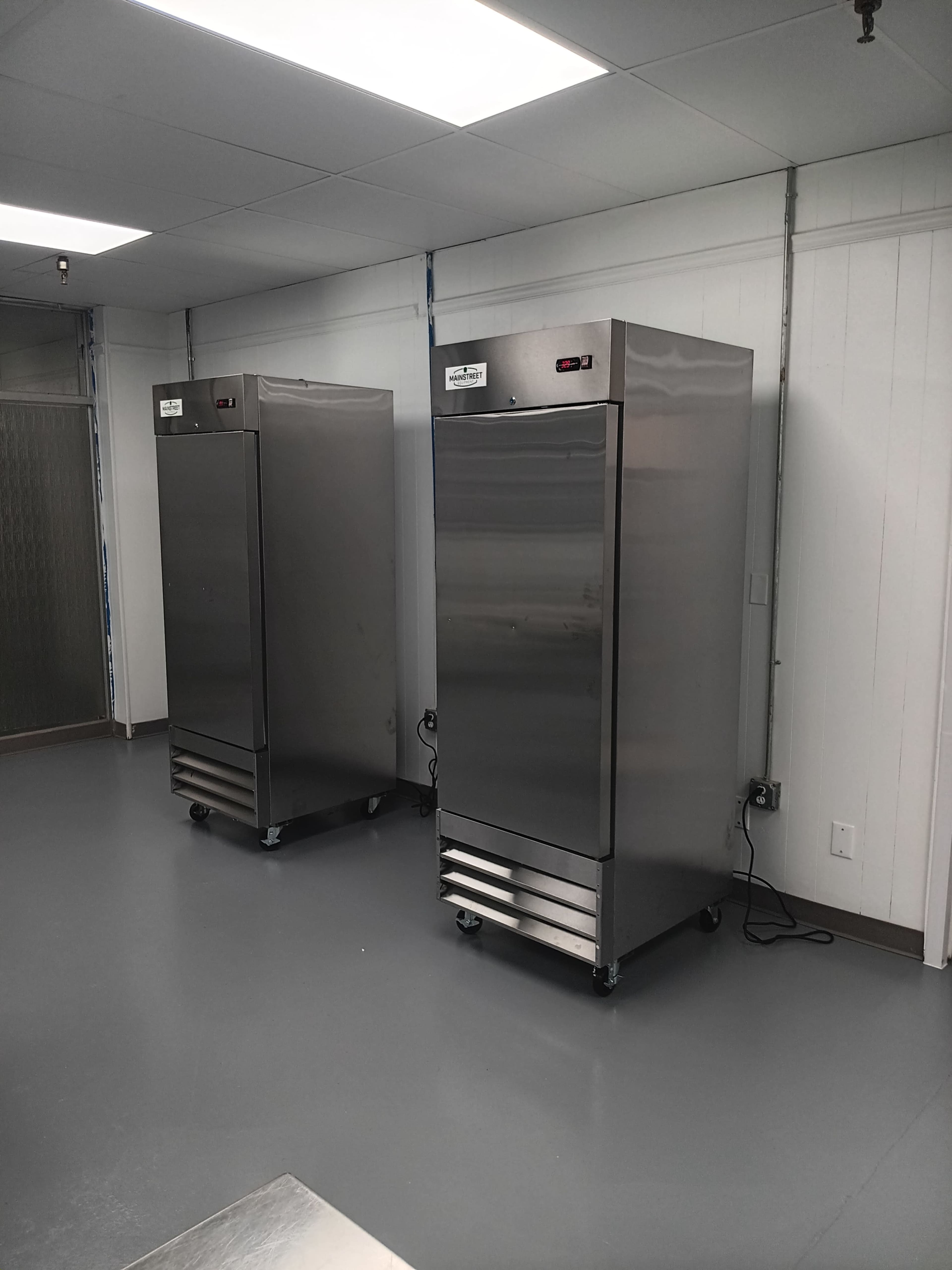 Two stainless steel commercial refrigerators stand side by side in a well-lit, empty kitchen space with a gray floor.