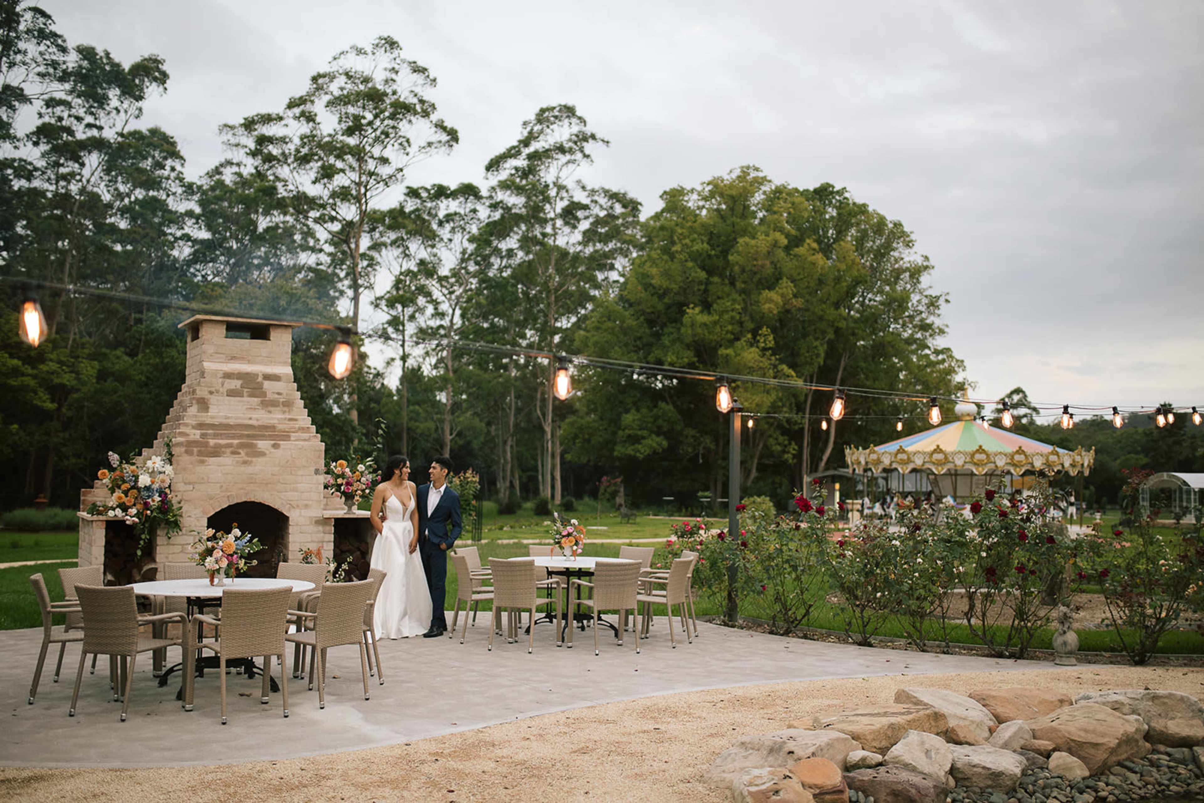 A couple stands together near a stone fireplace and outdoor seating area in a garden, with a carousel in the background.