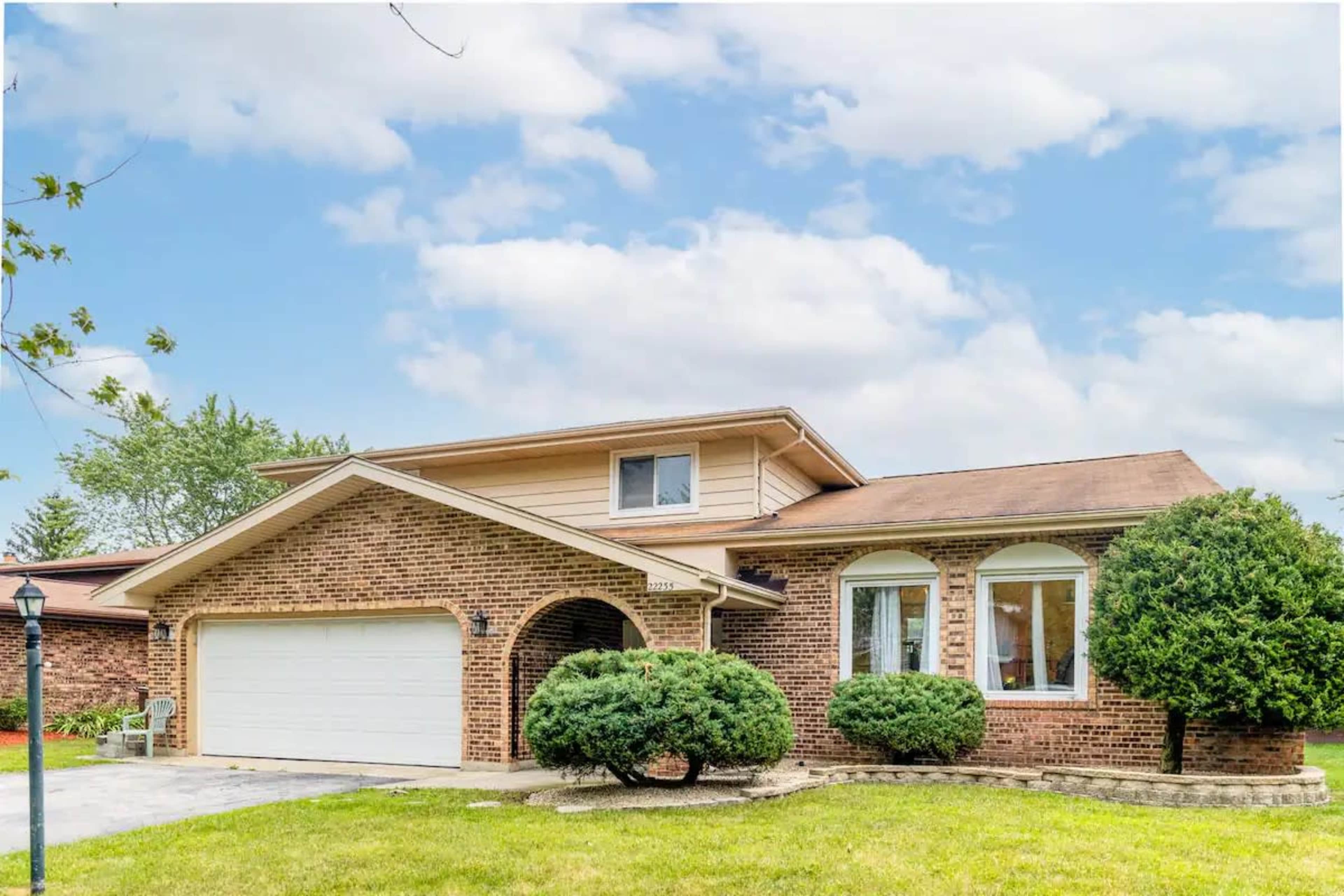 A two-story brick house with a garage and neatly trimmed bushes sits on a green lawn under a partly cloudy sky.