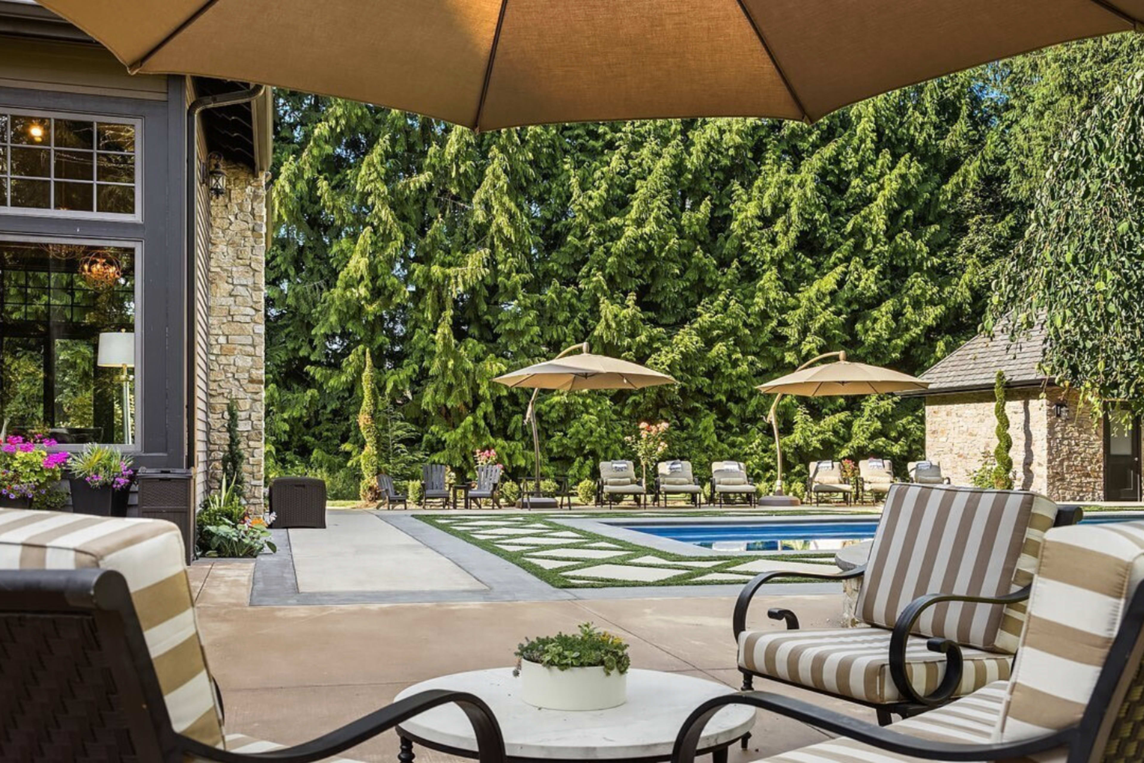 A patio area features striped lounge chairs, a round table with a plant, and a swimming pool surrounded by umbrellas and greenery.