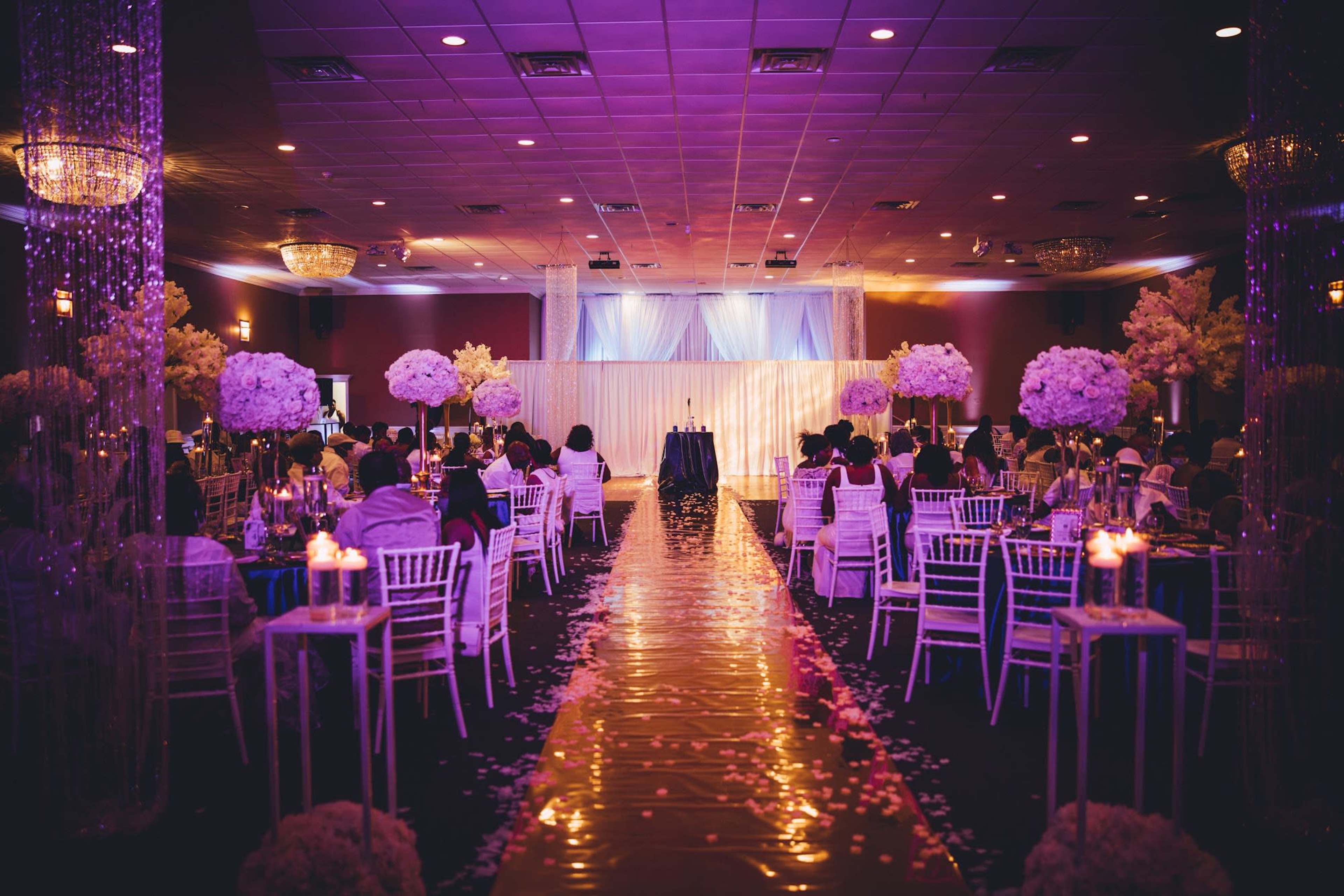 A decorated banquet hall with tables set for a formal event, featuring floral centerpieces and an aisle leading to a stage.