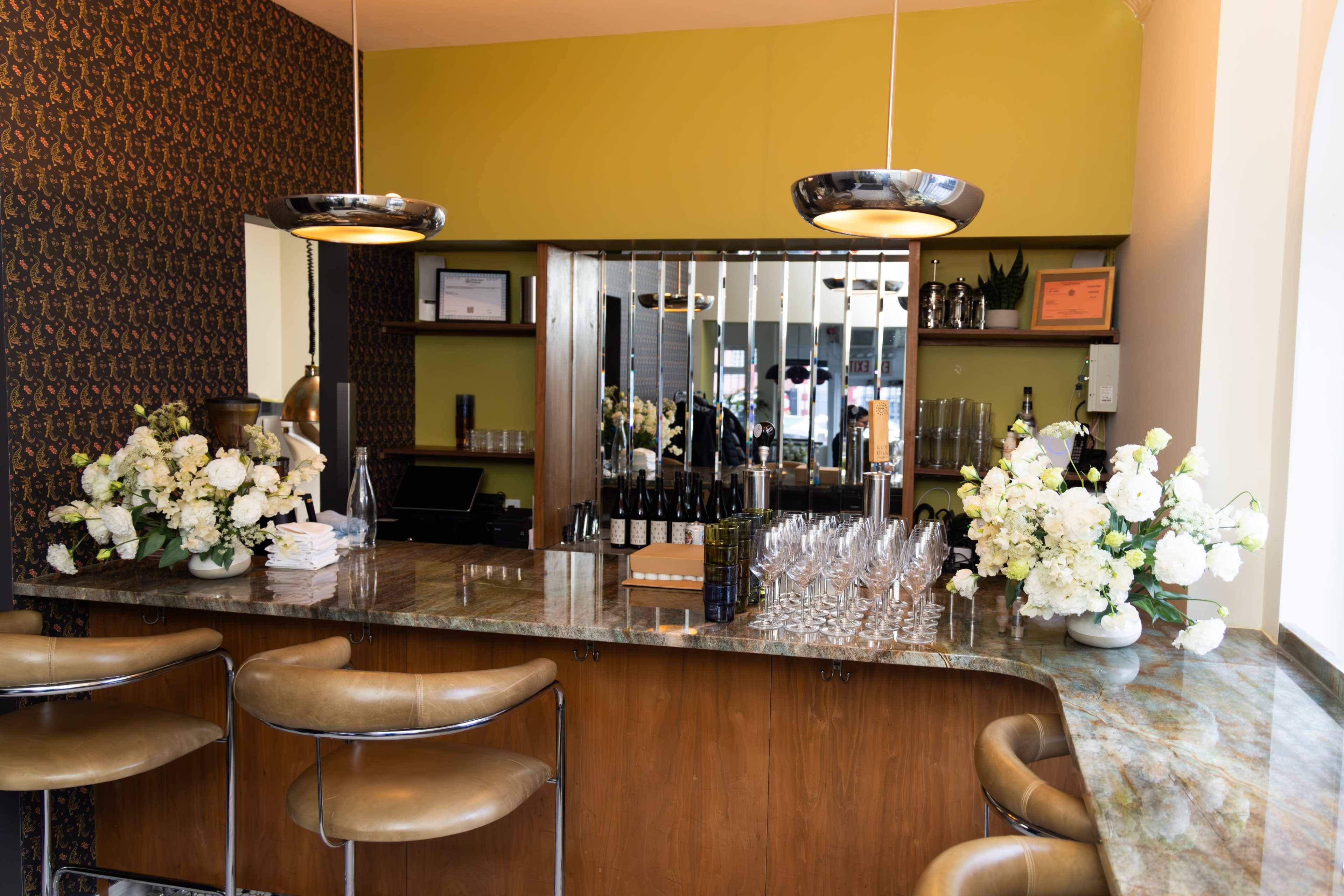 The image shows a stylish bar area with a marble countertop, modern pendant lighting, and floral arrangements on display.