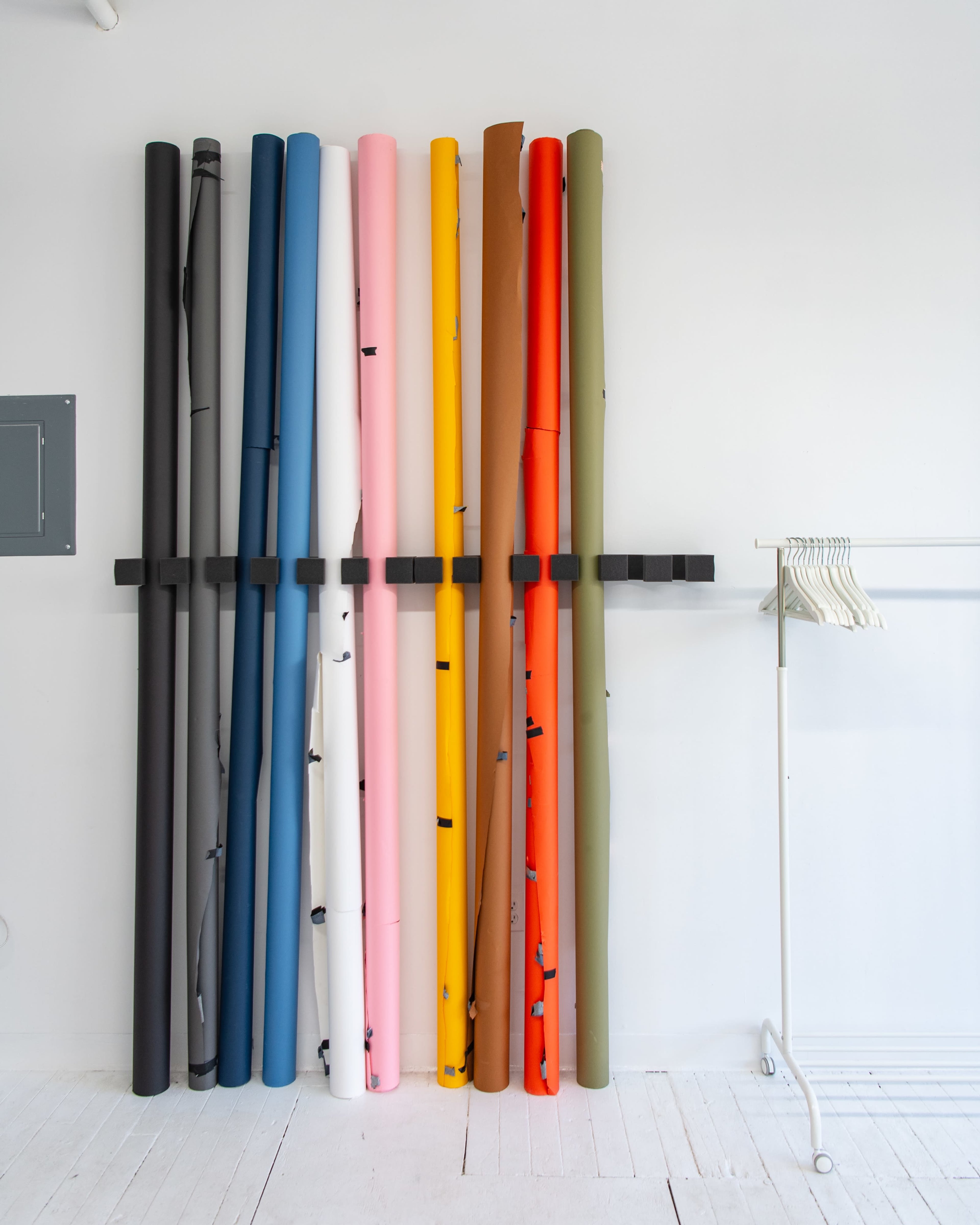 A series of colorful rolls of paper are neatly arranged vertically against a white wall, alongside a clothing rack with empty hangers.