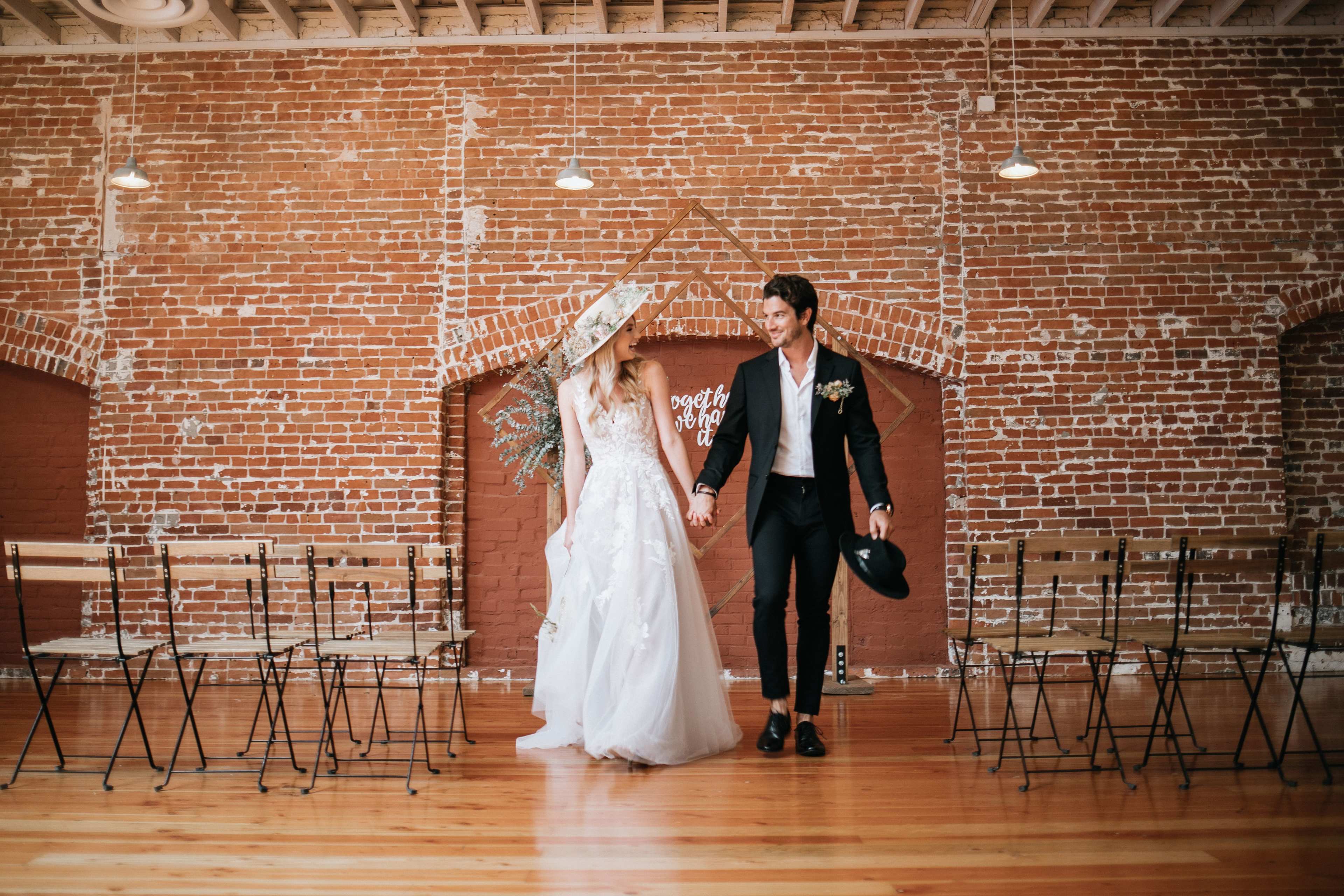 A couple in formal wedding attire walks hand in hand in front of a brick wall adorned with decorative elements, with metal chairs arranged for a ceremony.