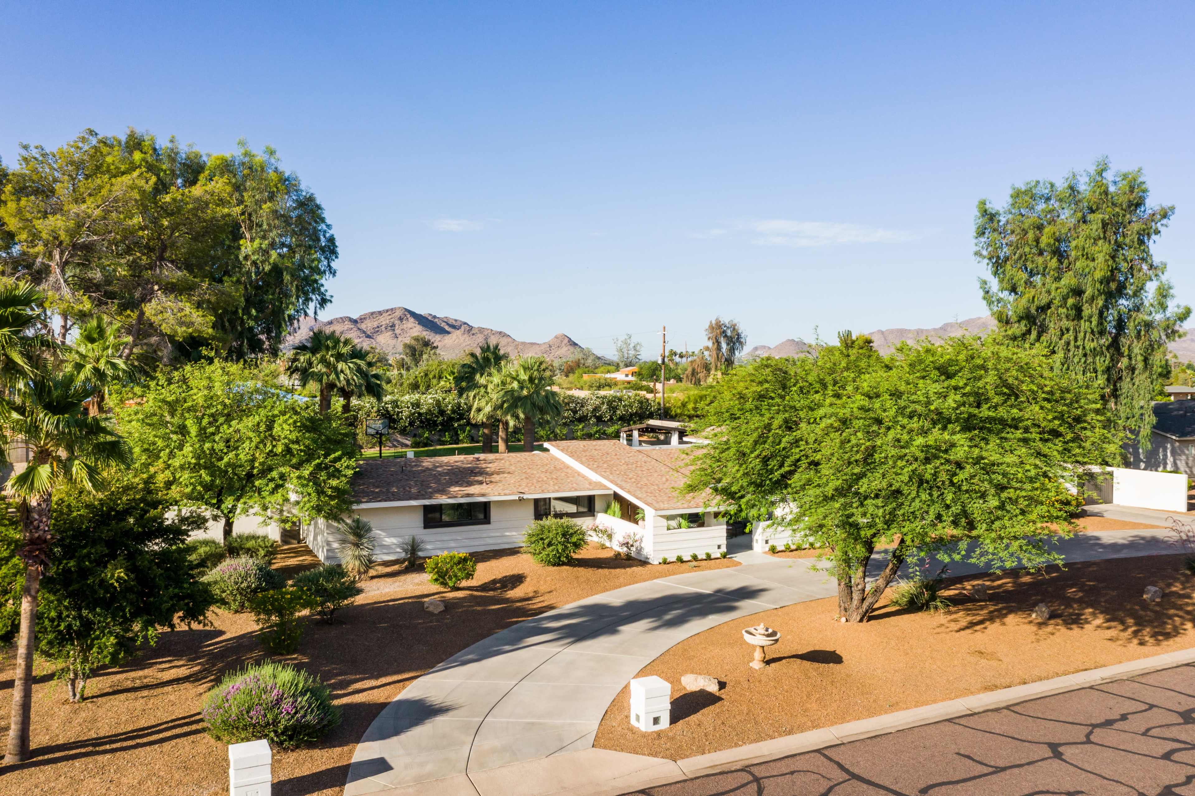 A single-story house with a circular driveway is surrounded by landscaped trees and mountains in the background.