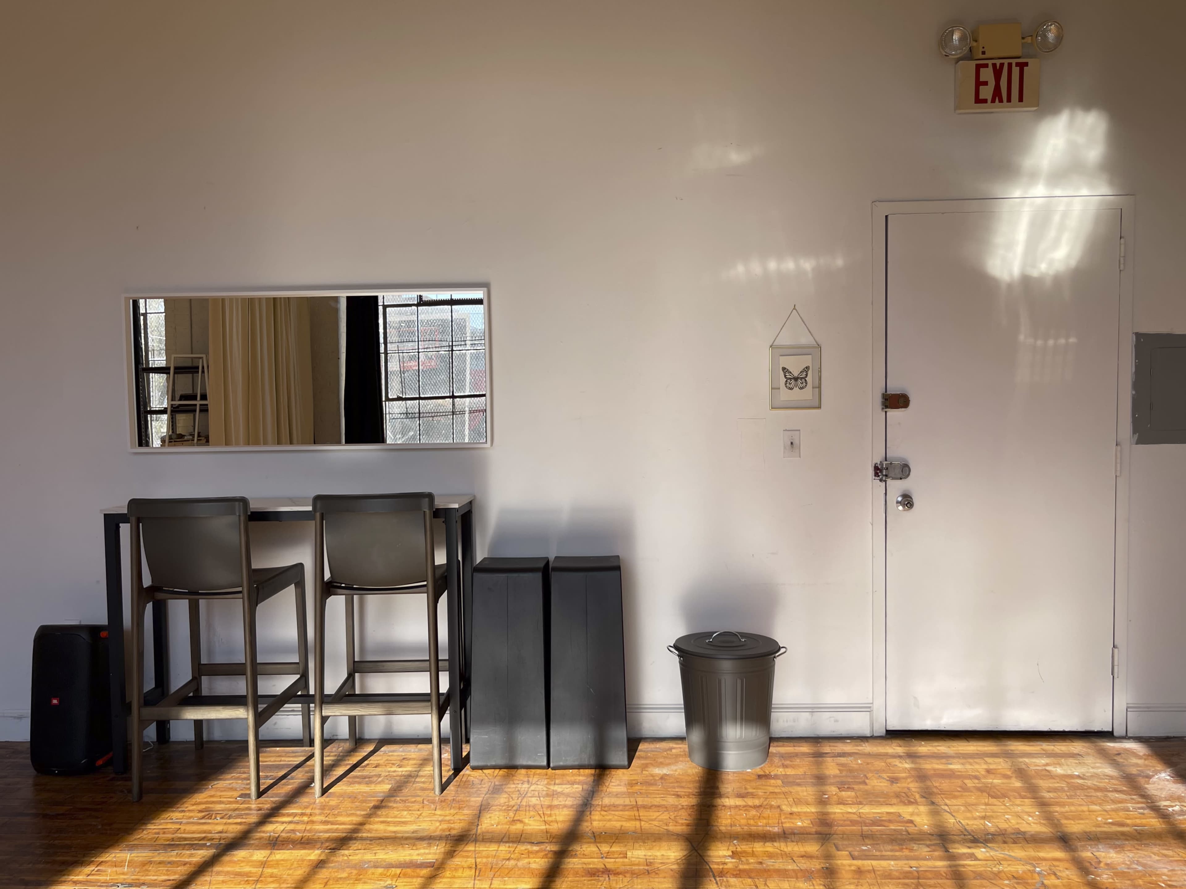 A minimalist interior space featuring a tall table with two chairs, a mirror, a door labeled "EXIT," two stacked storage bins, and a trash can on the floor.