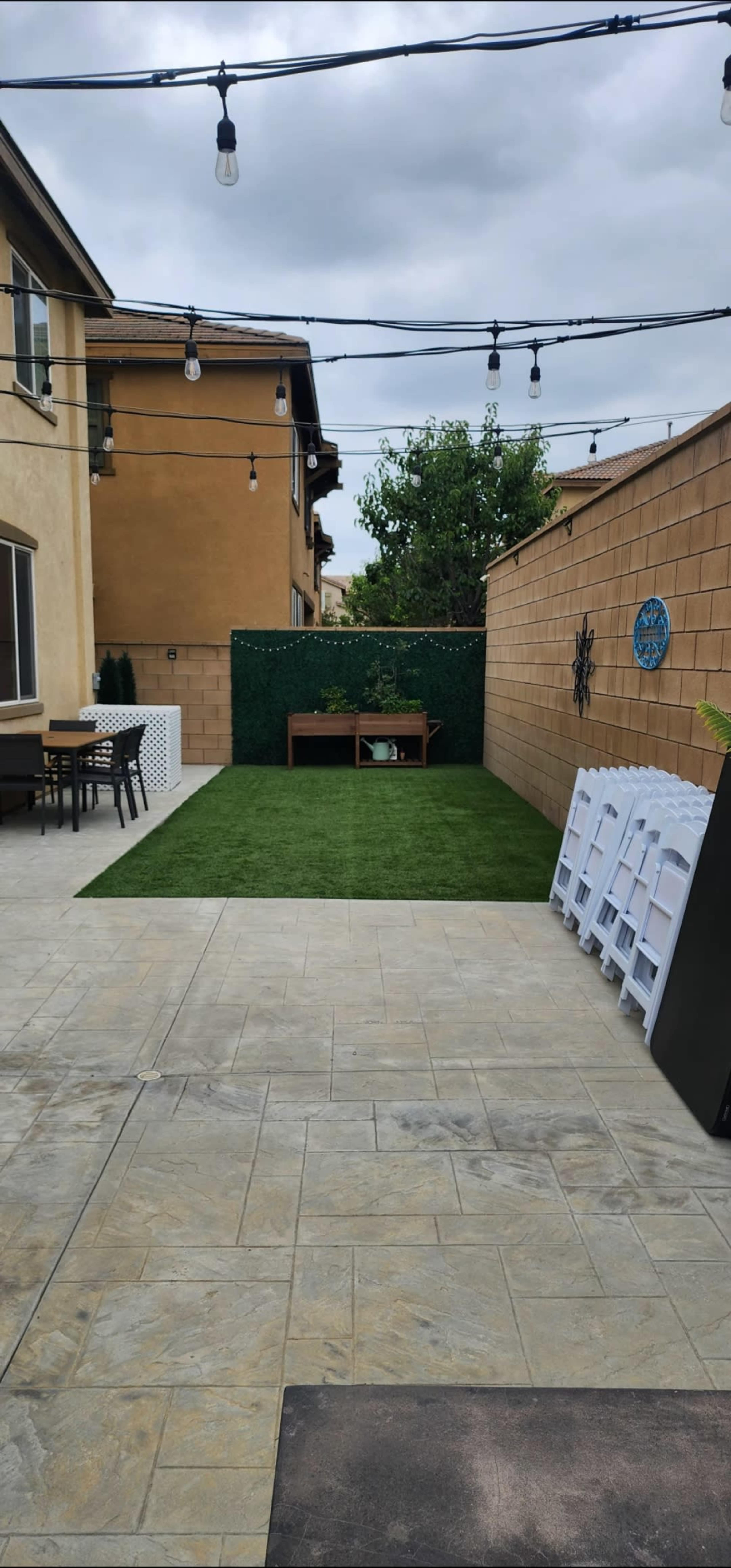 The image shows a paved backyard with artificial grass, a wooden table set with chairs, and string lights overhead.