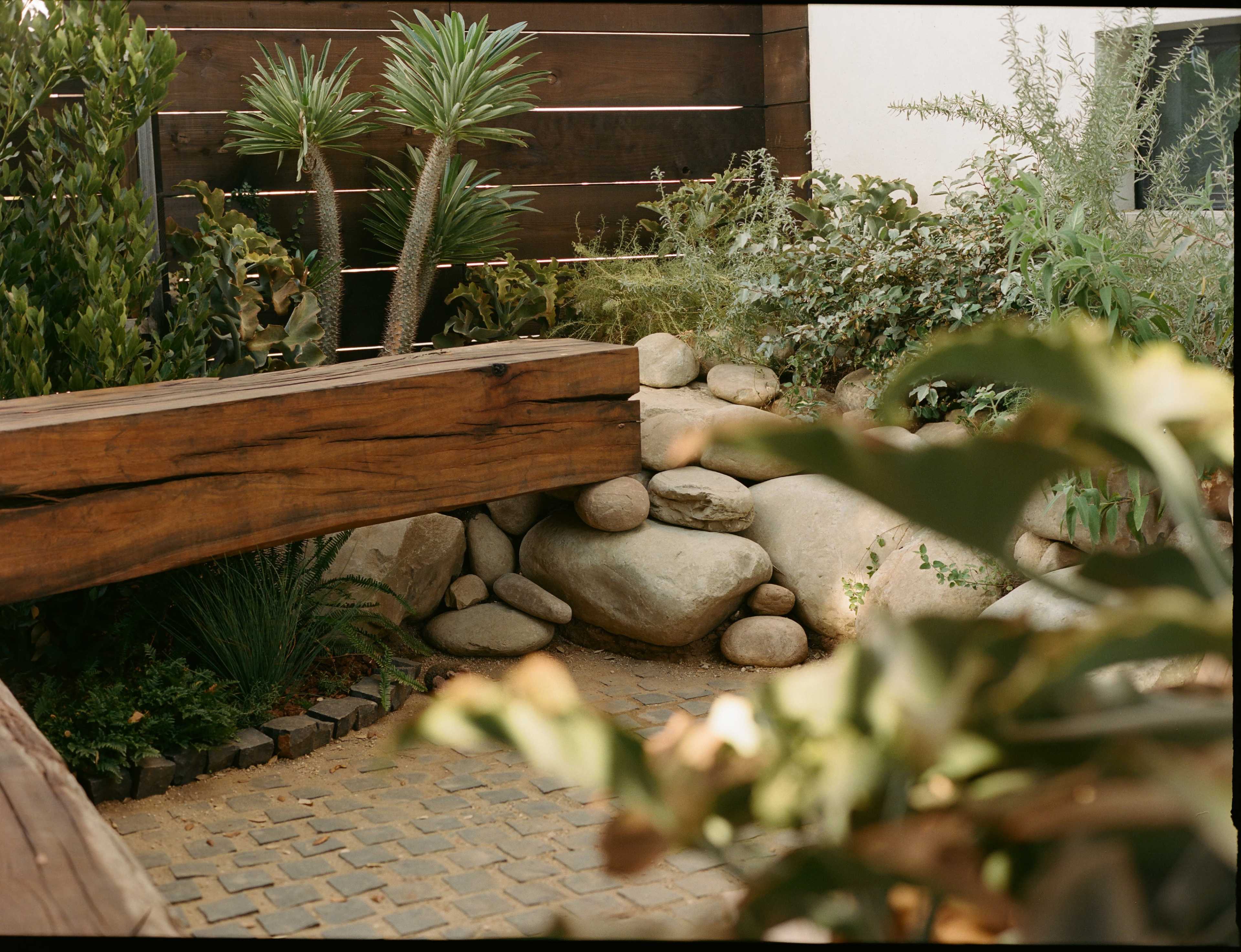 A landscaped area featuring a wooden bench, smooth stones, and various plants enclosed by a wooden fence.