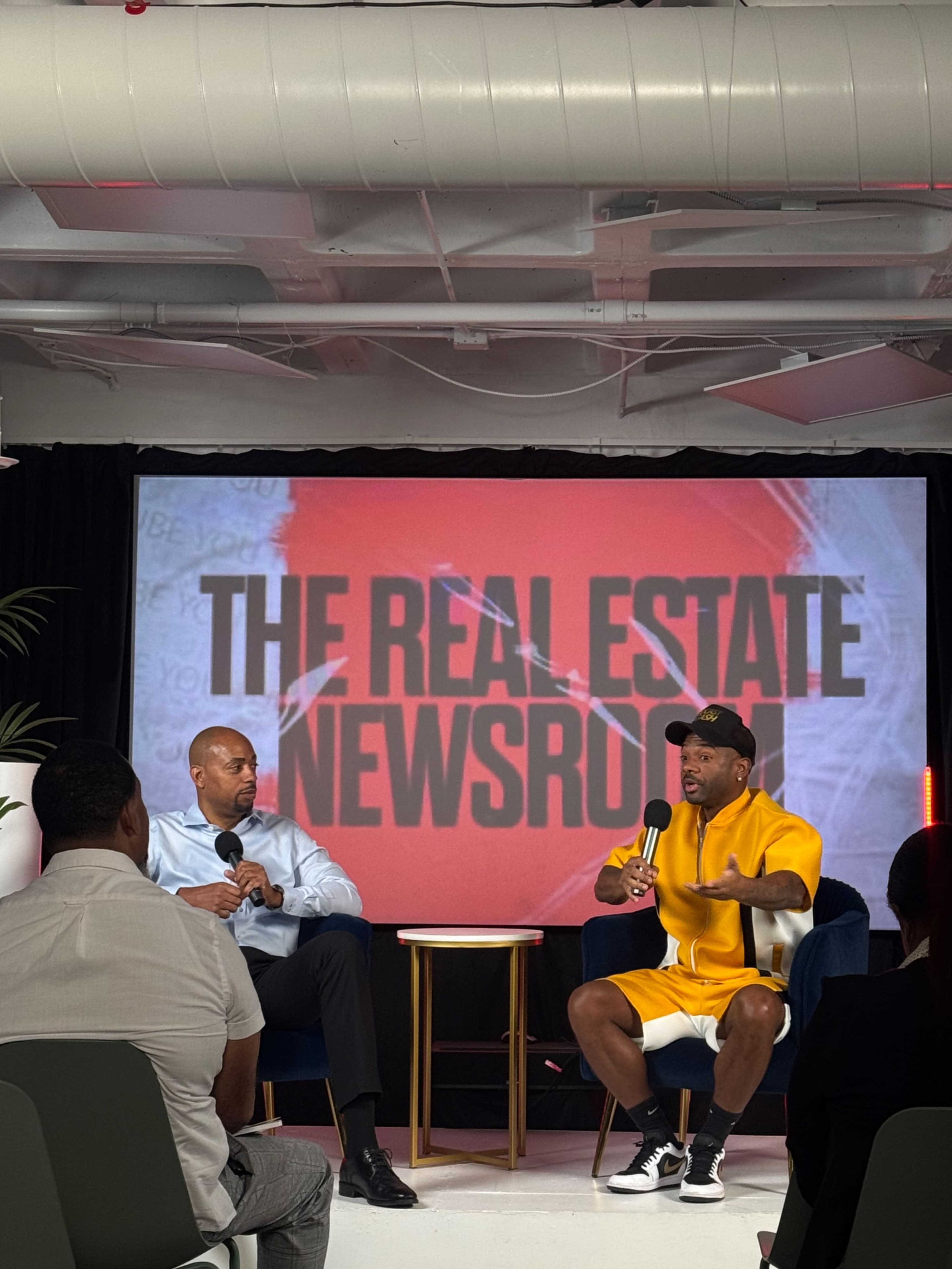 A man in a bright yellow outfit speaks while seated on a stage, as another man listens attentively nearby, with a large screen behind displaying "THE REAL ESTATE NEWSROOM."