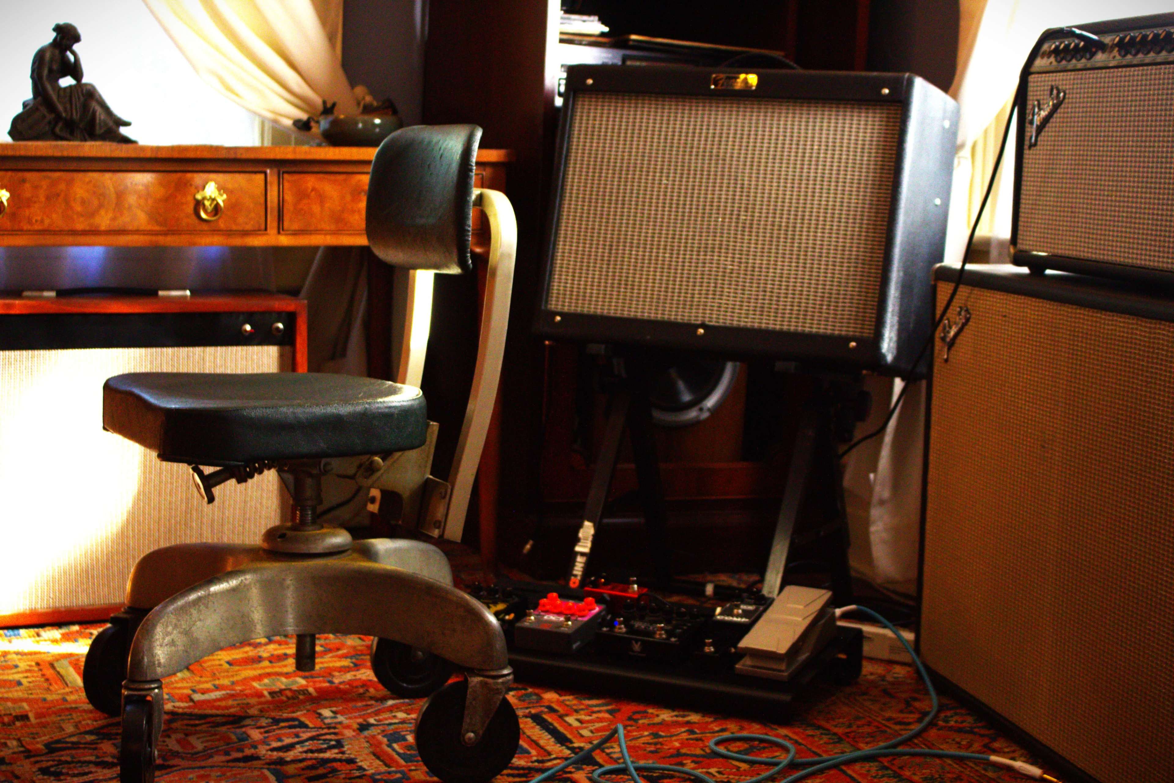 A black office chair is positioned next to a guitar amplifier and effects pedals, surrounded by wooden furniture and textured rugs.