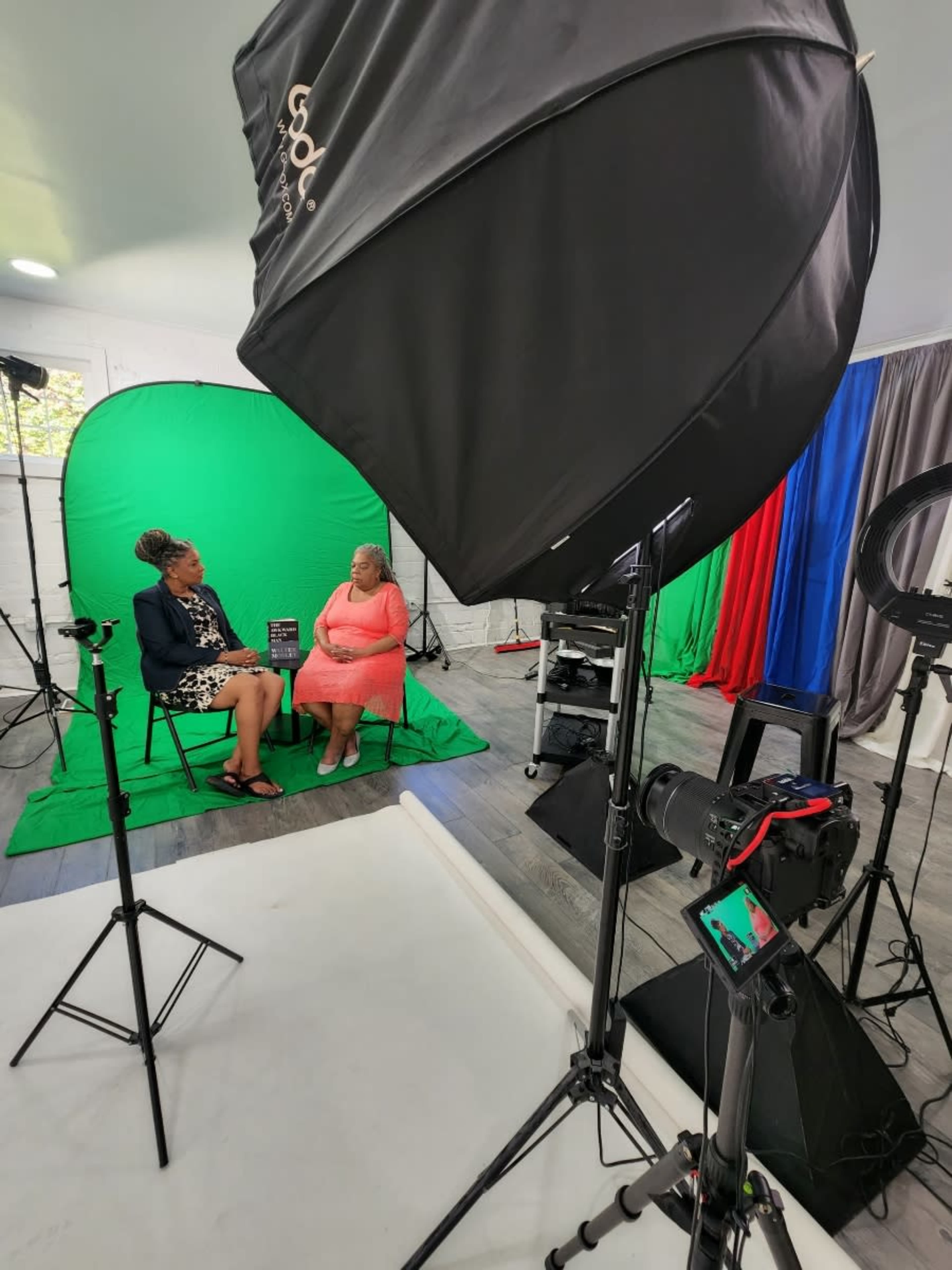 Two women are seated for an interview in a studio with green and red backdrops, surrounded by lighting equipment and cameras.