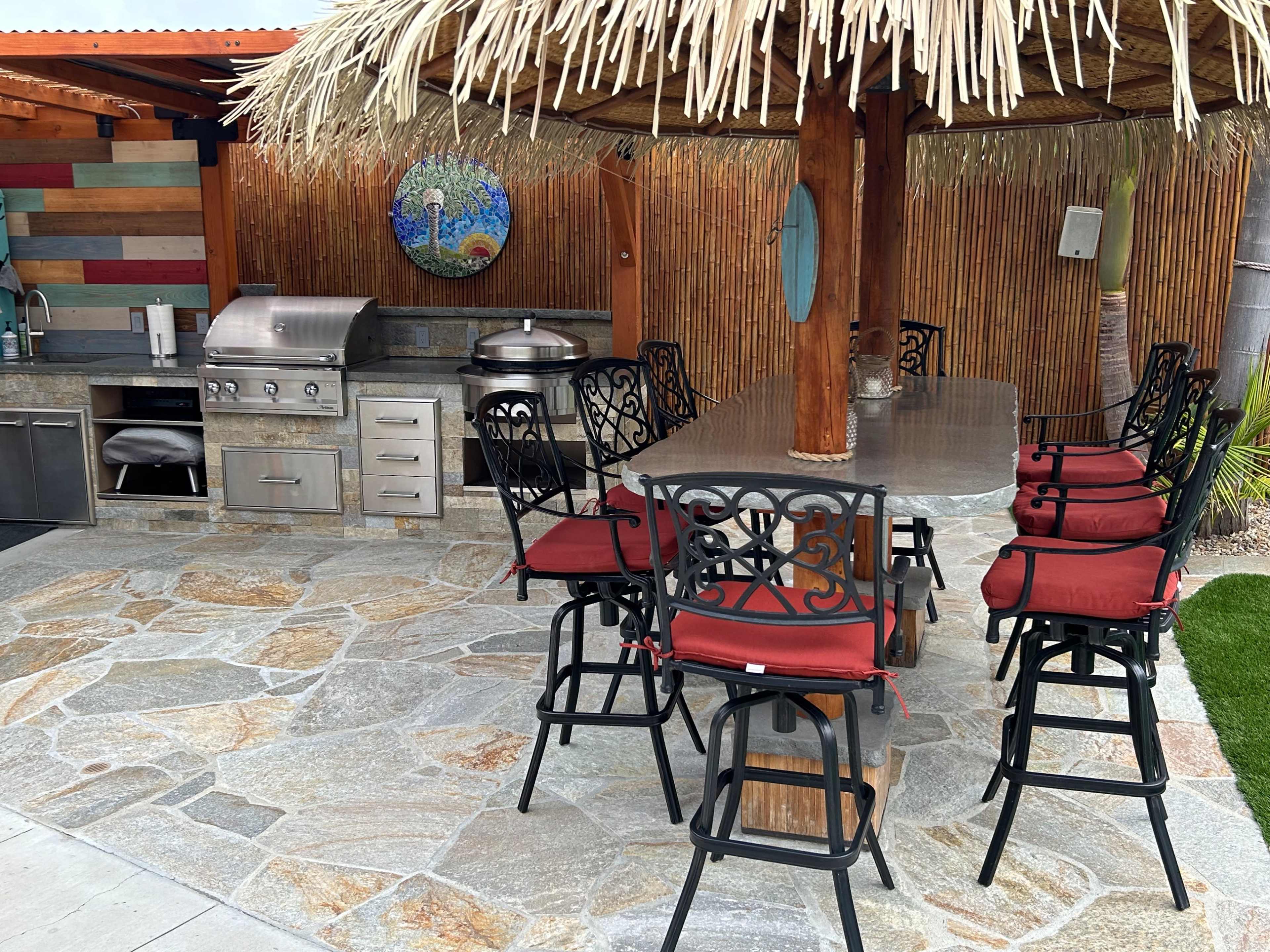 An outdoor kitchen area with a stone floor, a large barbecue grill, and a bar with high stools under a thatched roof.