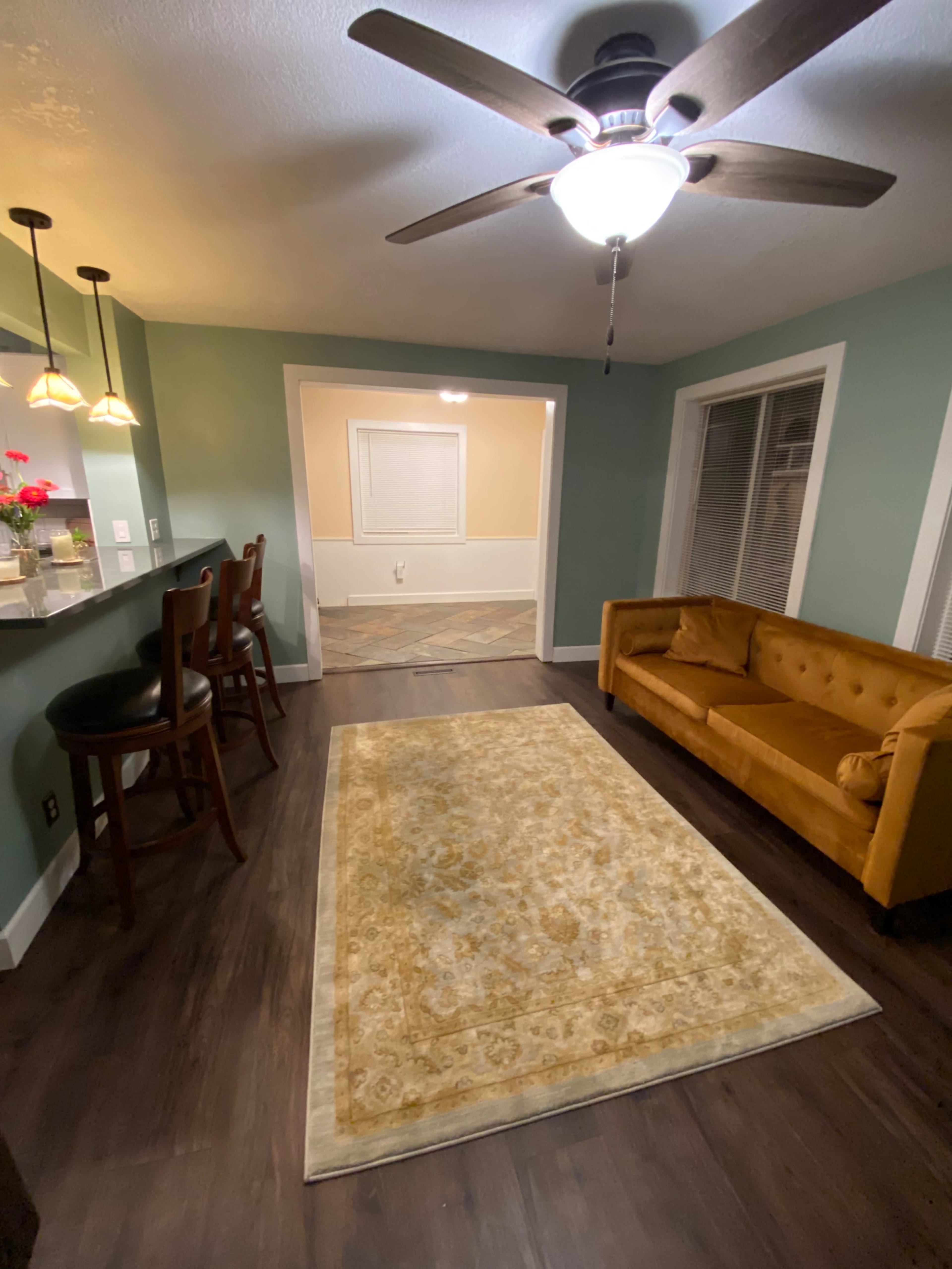 The image shows a living room with a yellow couch, a decorative rug, and a bar area with three stools against a green wall.