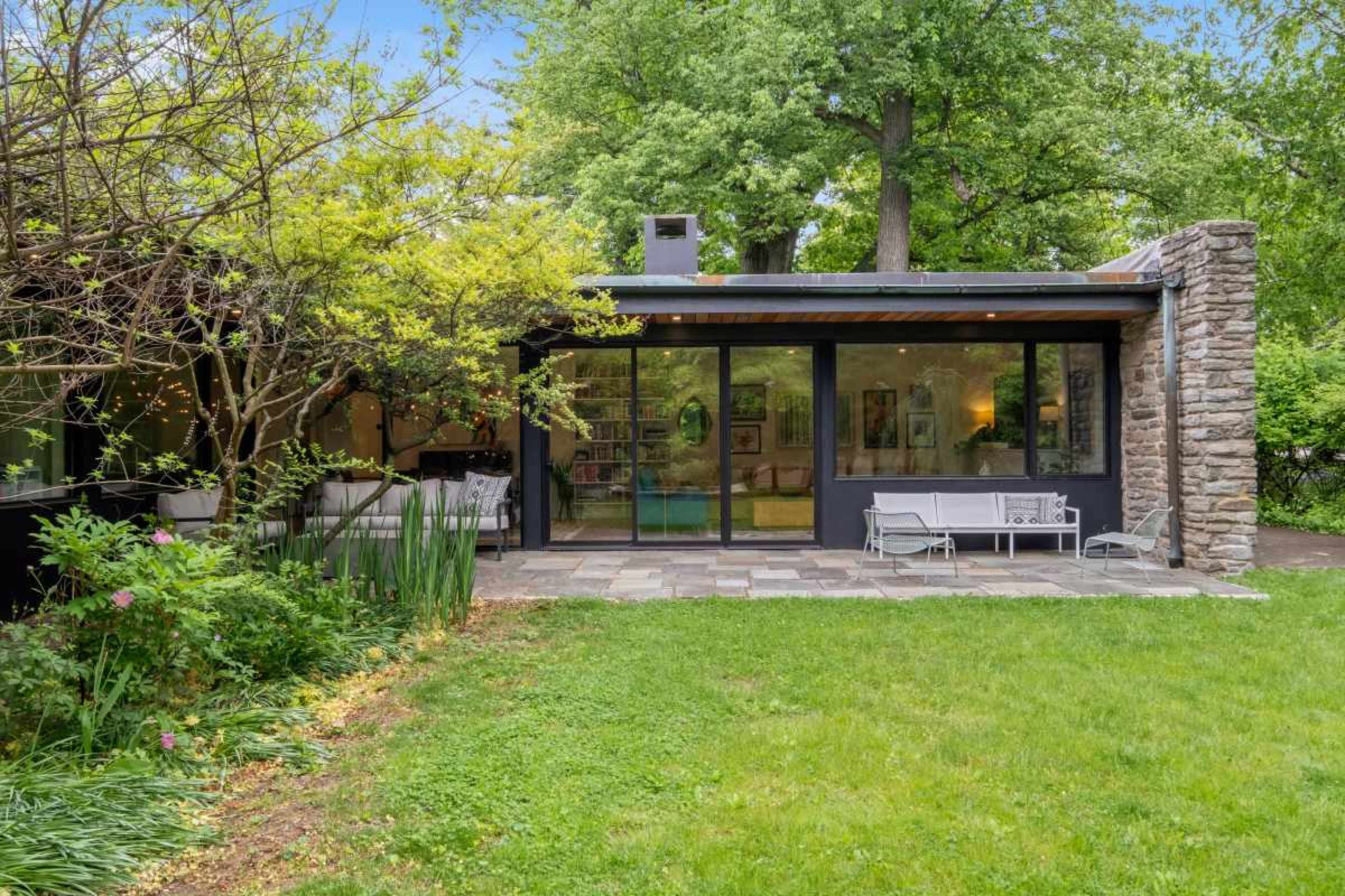 The image shows a modern home with a stone exterior and large glass windows, set in a lush green garden with trees and plants.