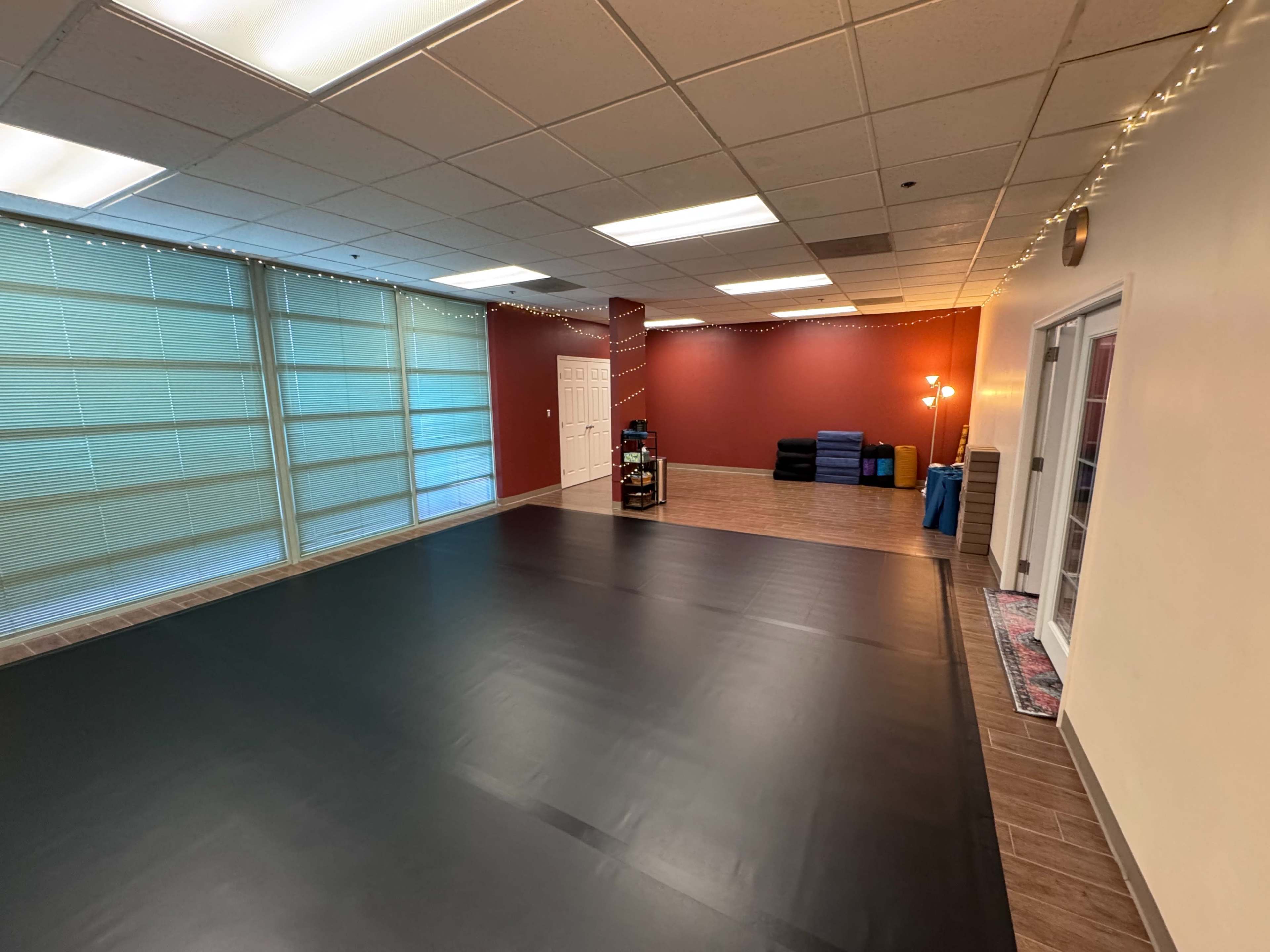 The image shows a spacious room with a black mat on the floor, light-colored walls, and windows lined with sheer curtains.