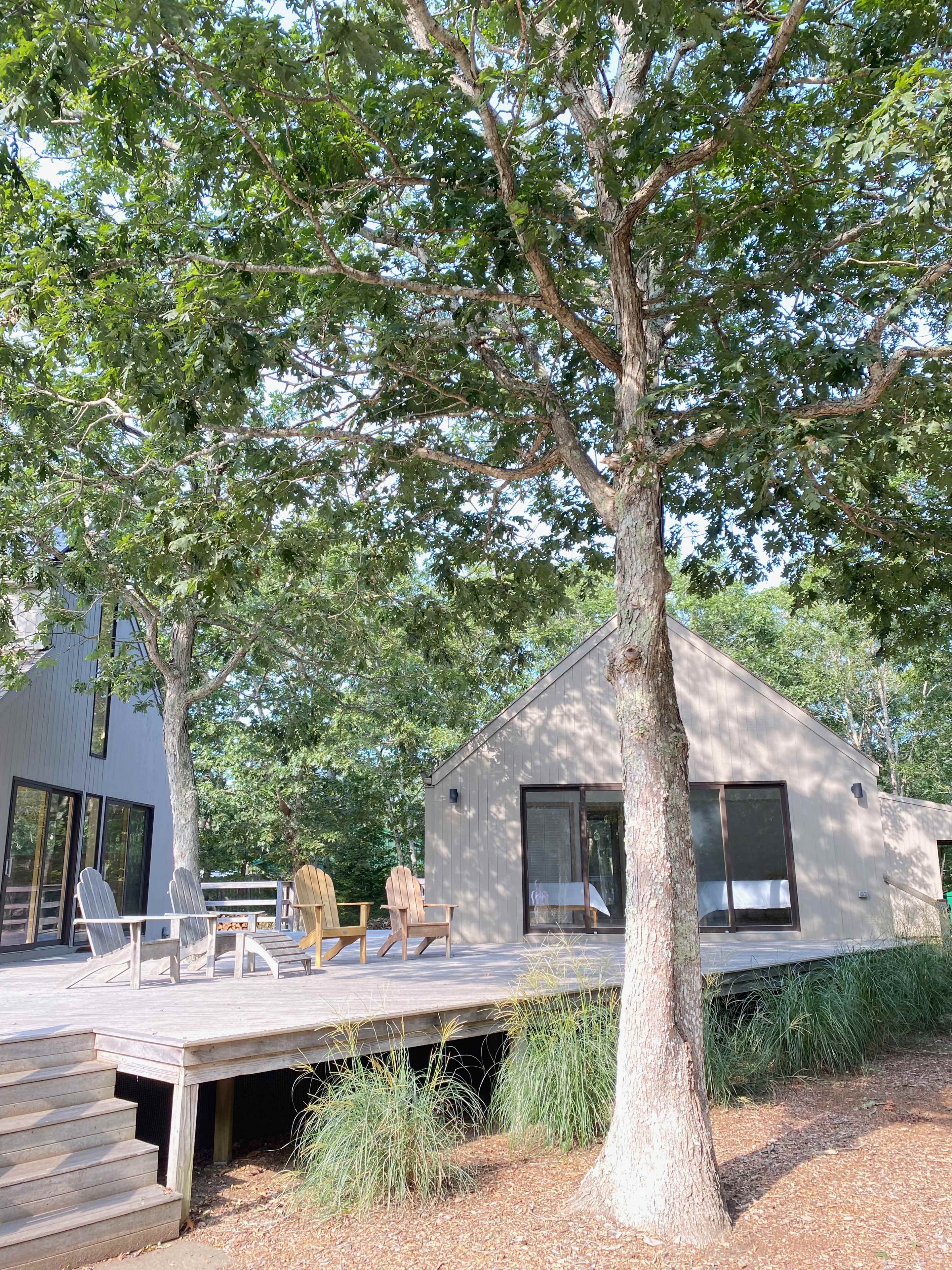 A modern-style house with large windows is surrounded by trees and features a wooden deck with several chairs.