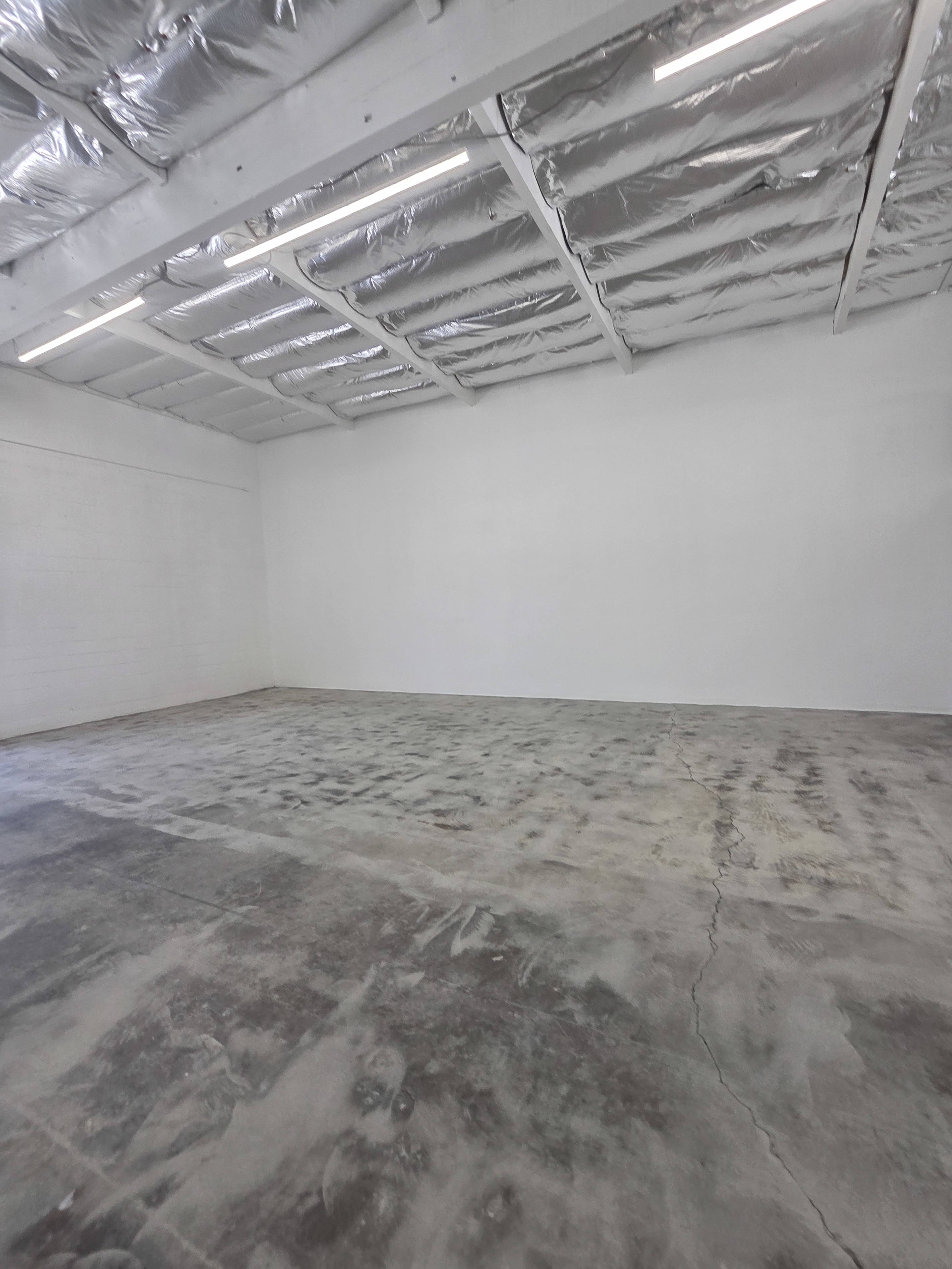 The image shows an empty, spacious room with a concrete floor and white walls, illuminated by overhead lights.