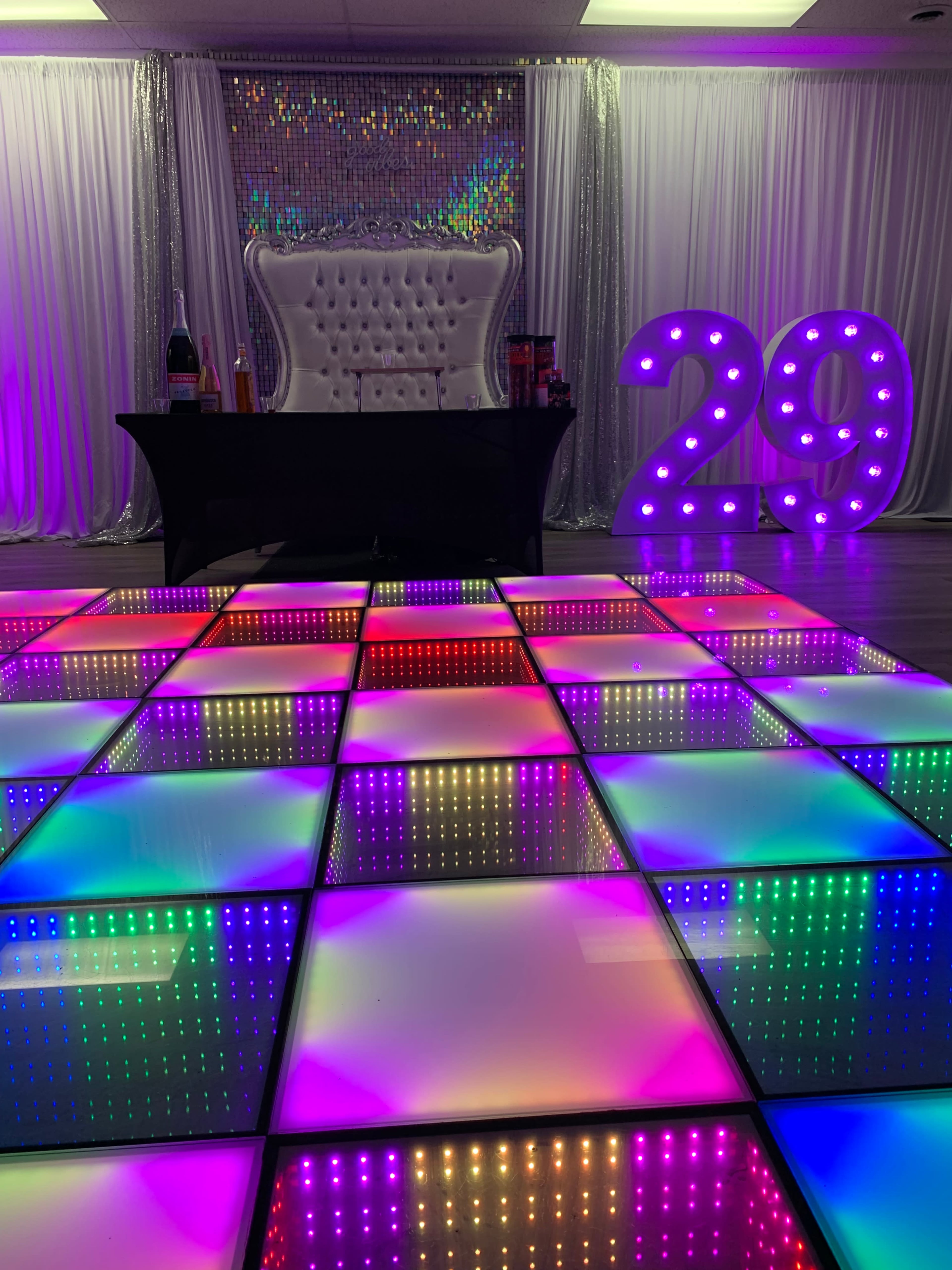 The scene features a vibrant event space with a illuminated dance floor, a black table, a white tufted backdrop, and a large number "29" displayed in lights.