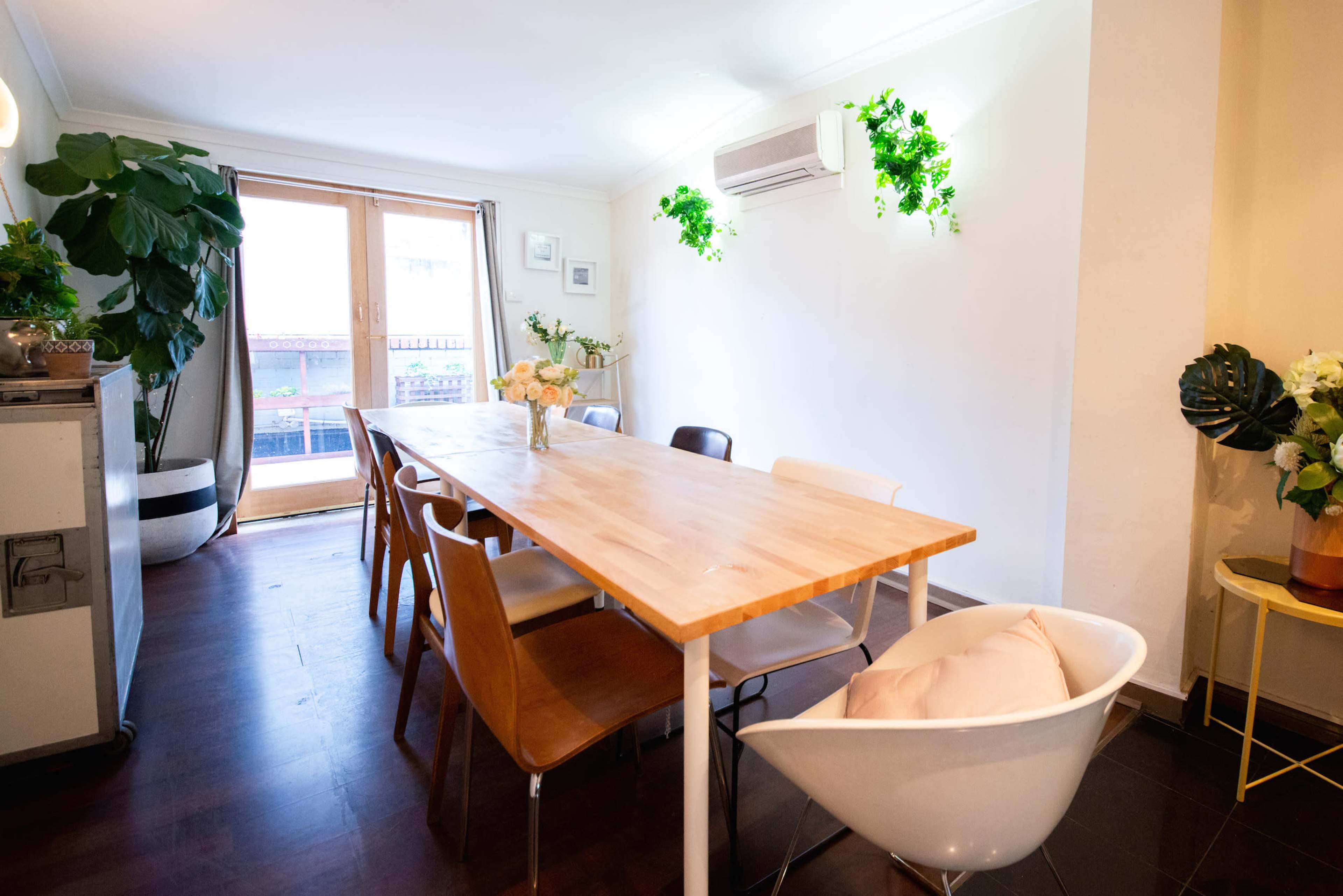 A large wooden dining table is surrounded by various chairs in a well-lit room with potted plants and an air conditioning unit mounted on the wall.