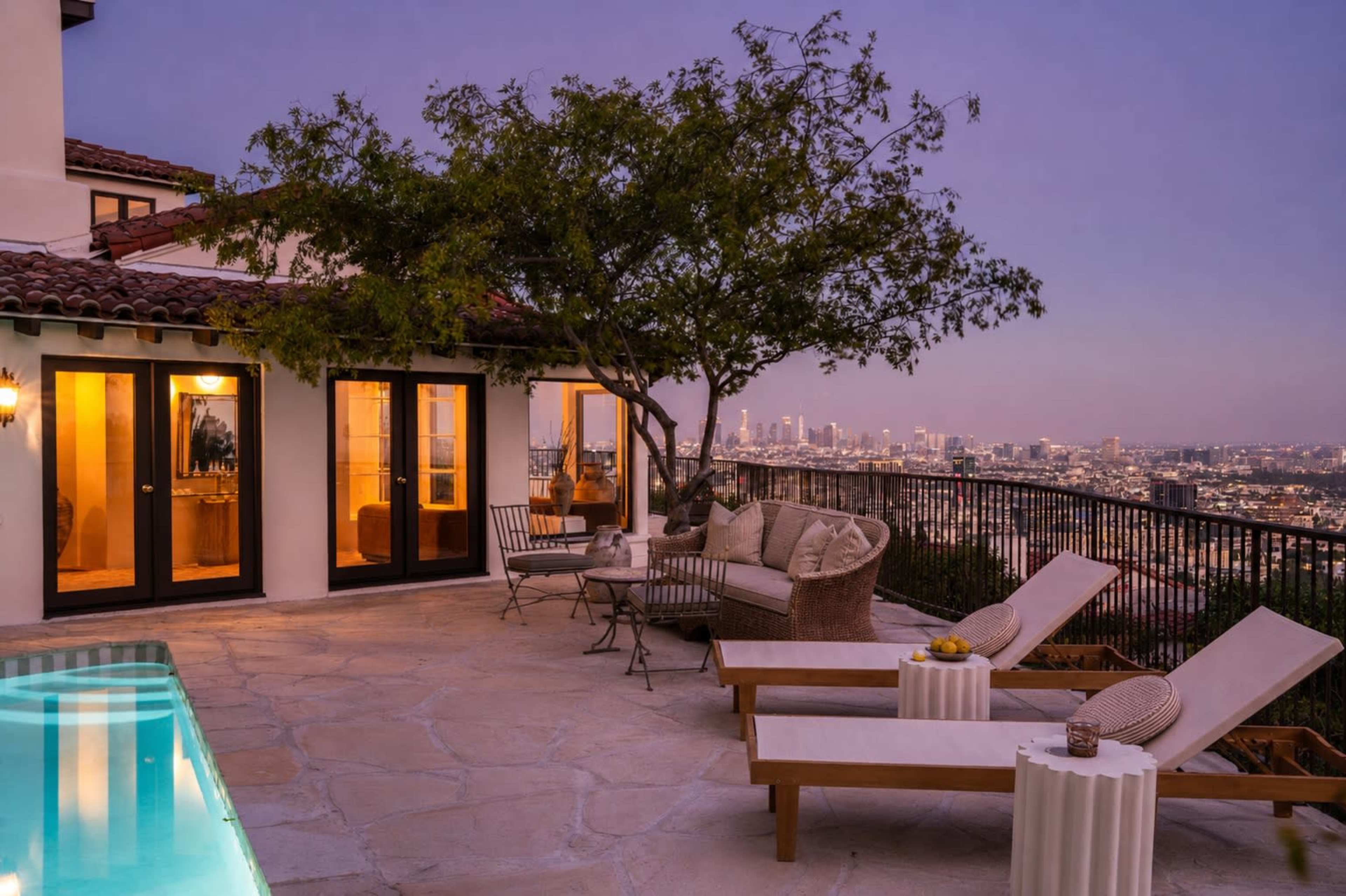 The scene shows a terrace with lounge chairs and a small table overlooking a city skyline at dusk.
