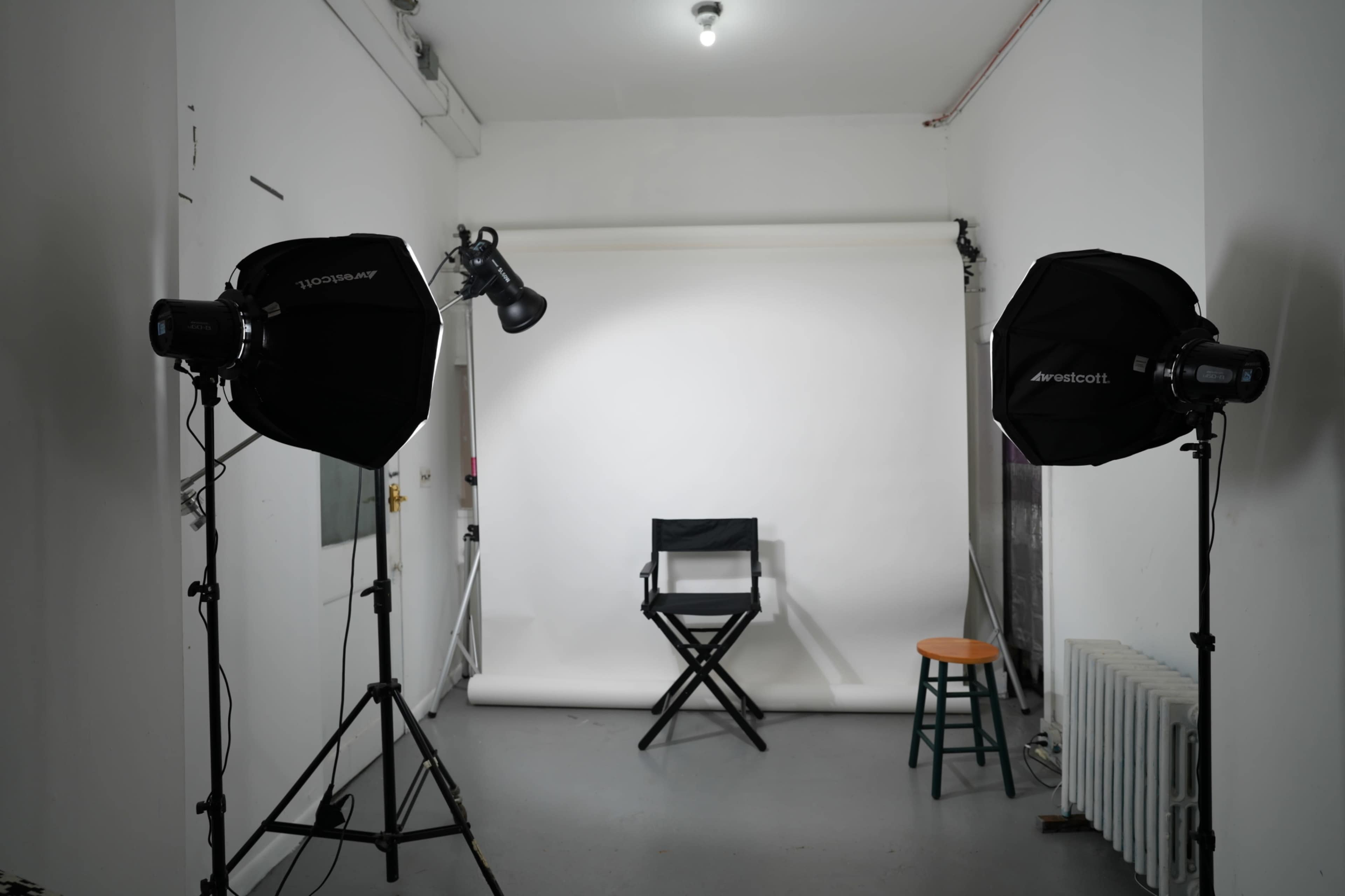 A photography studio features a white backdrop, a director's chair, and lighting equipment on tripods.