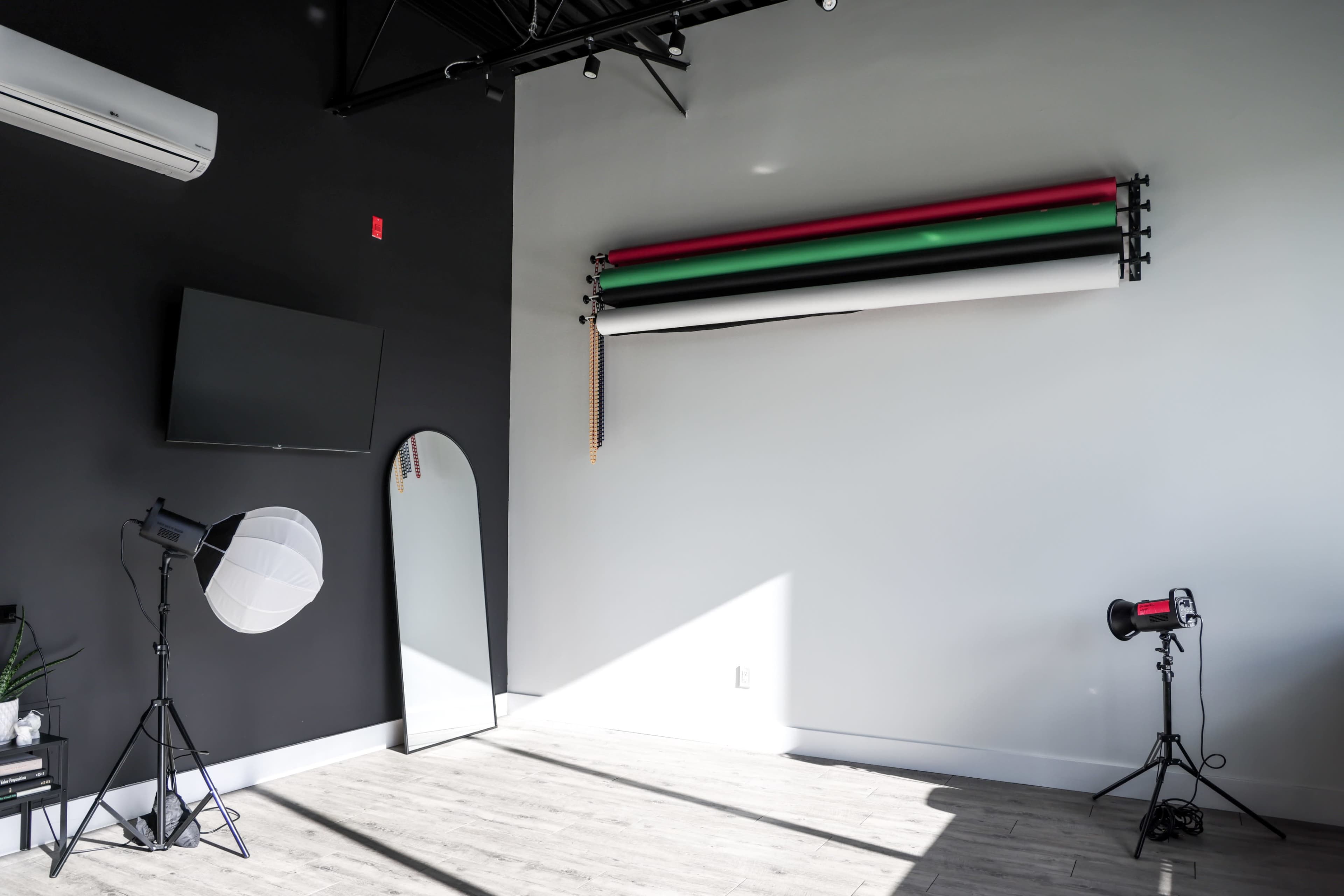 The image features a photography studio with black and white walls, a large mirror, a television, and rolls of backdrop paper in red, green, and black mounted on the wall.