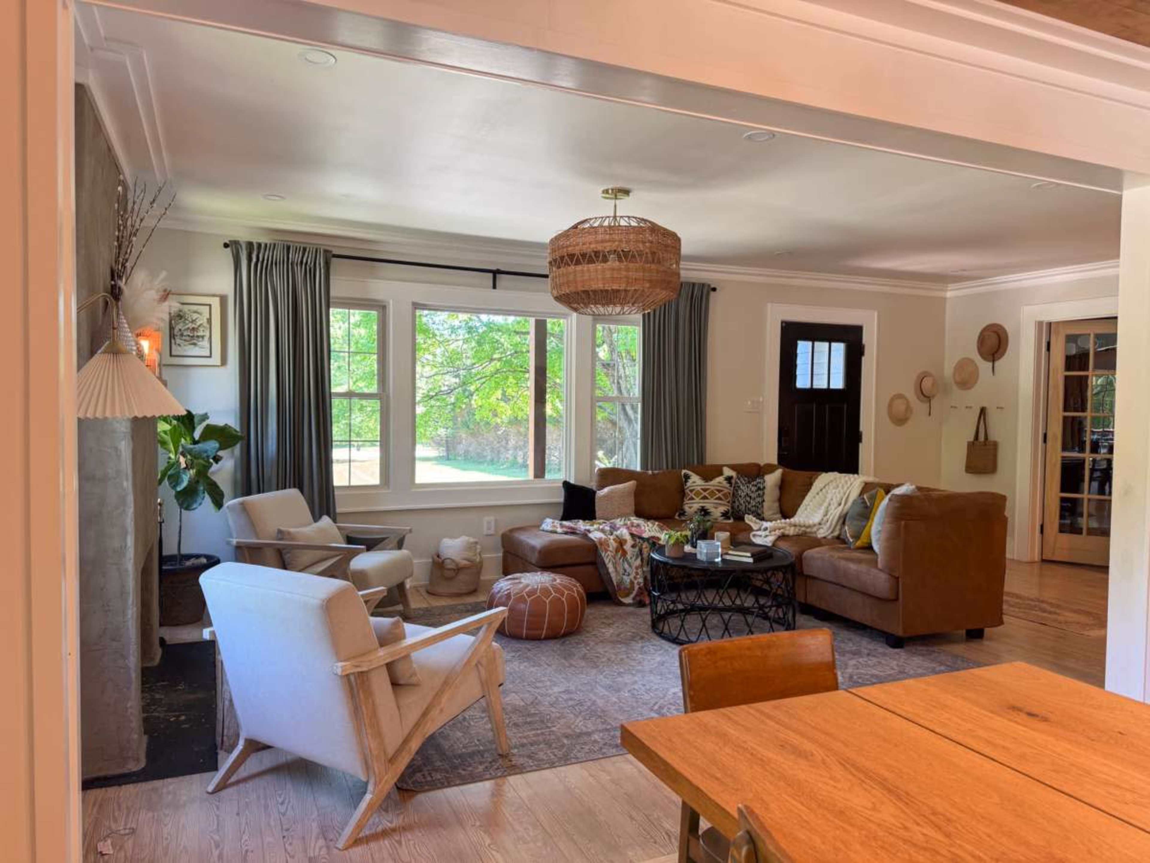 The image shows a cozy living room with a combination of brown leather and upholstered furniture, a large window providing natural light, and a variety of decorative items on the walls and furniture.