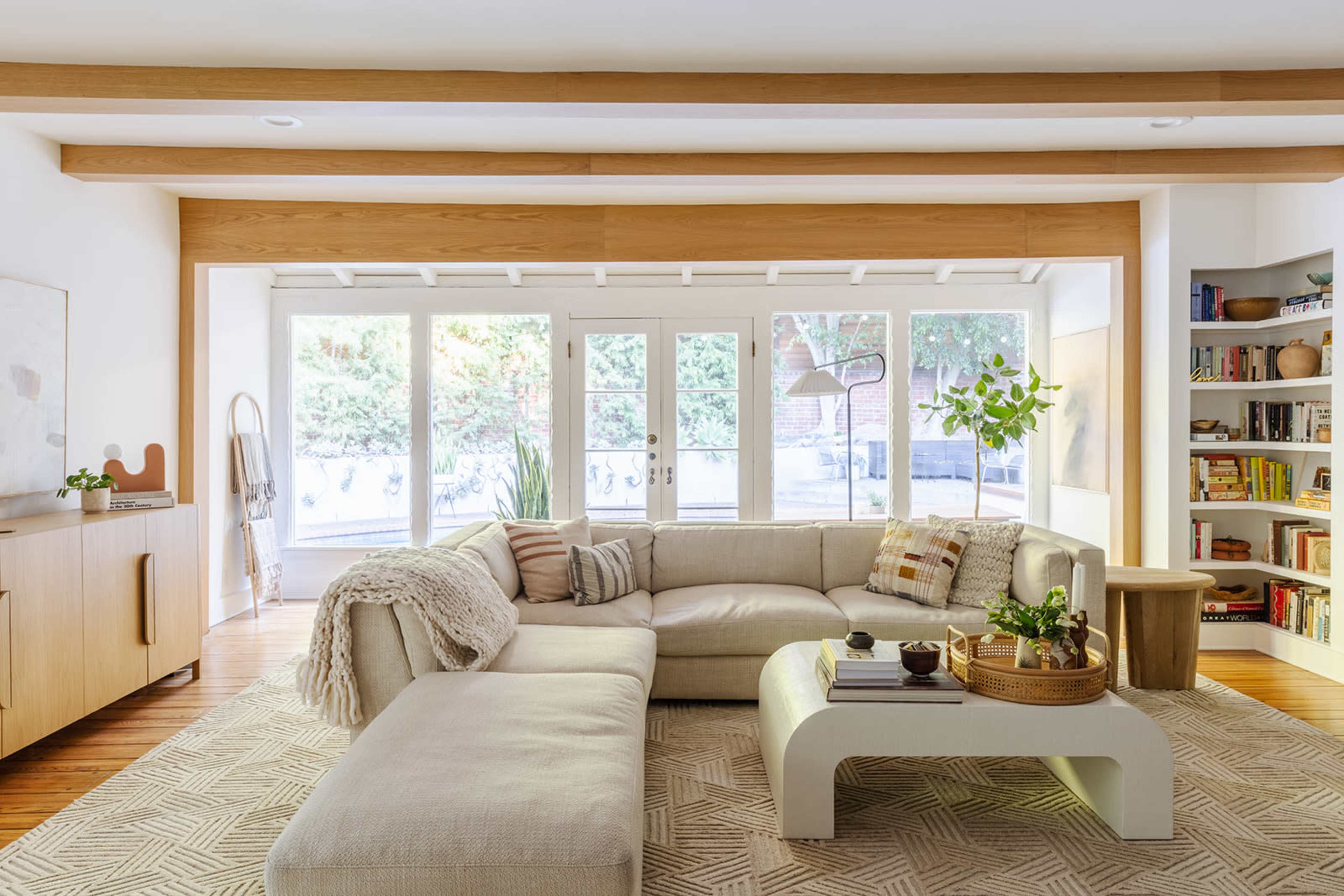 A light-filled living room features a large, cream-colored sectional sofa, a wooden coffee table, and a bookshelf filled with books.