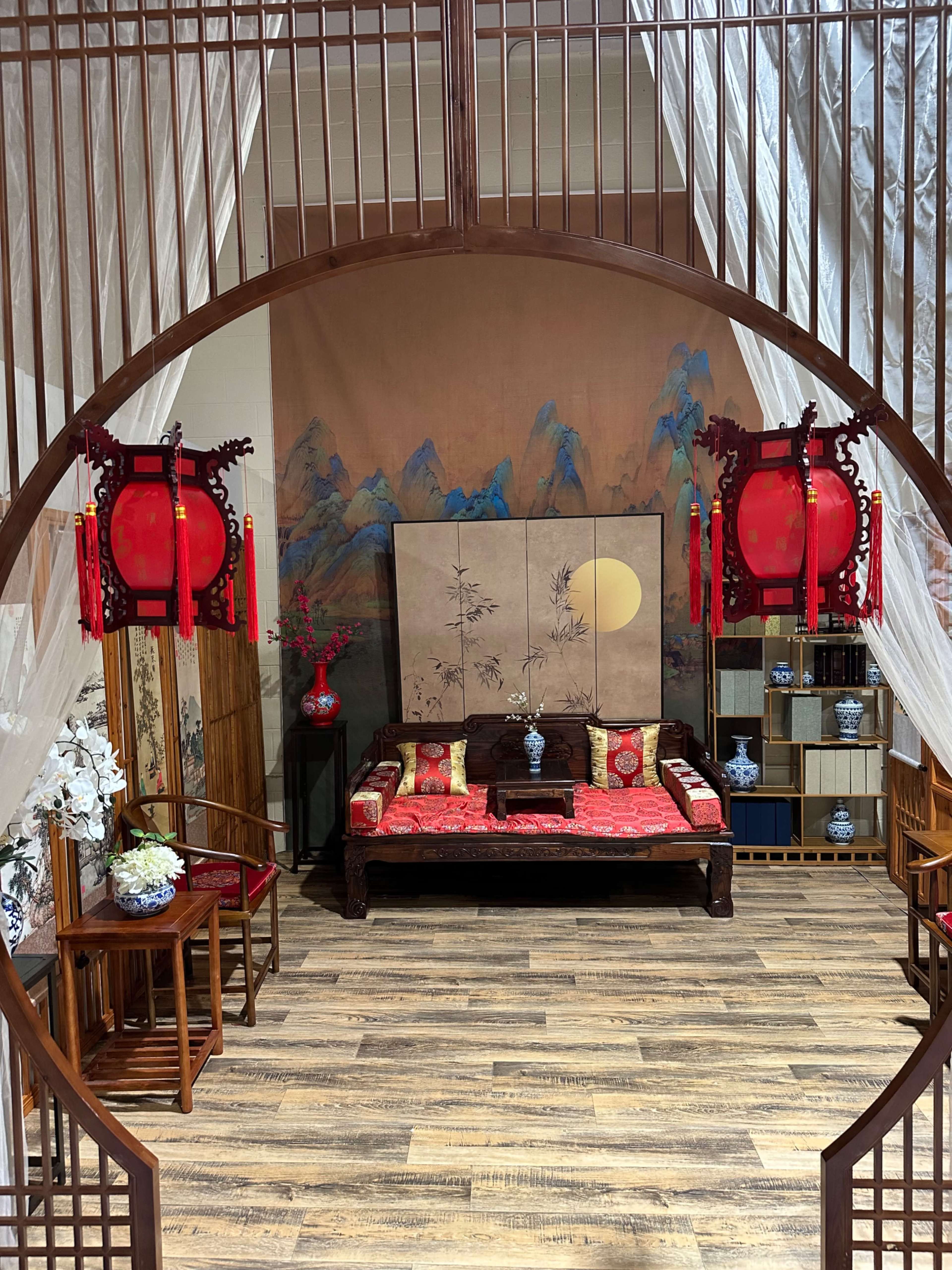 The image depicts a traditional Chinese interior with a circular entrance, red lanterns, a wooden sofa adorned with cushions, and decorative elements including a large painting and potted plants.