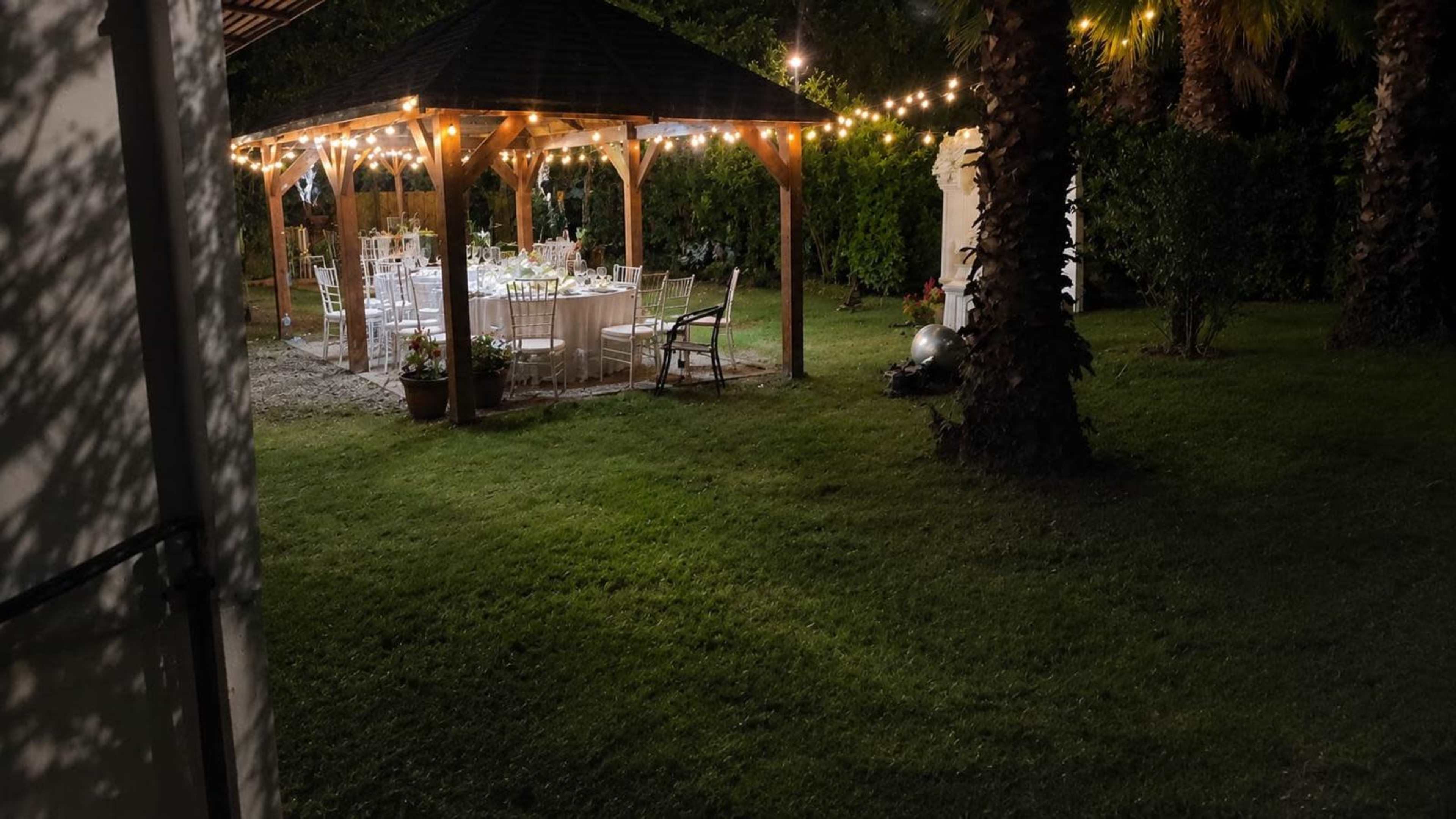 A decorated gazebo with a dining setup and string lights is located in a grassy backyard surrounded by trees.
