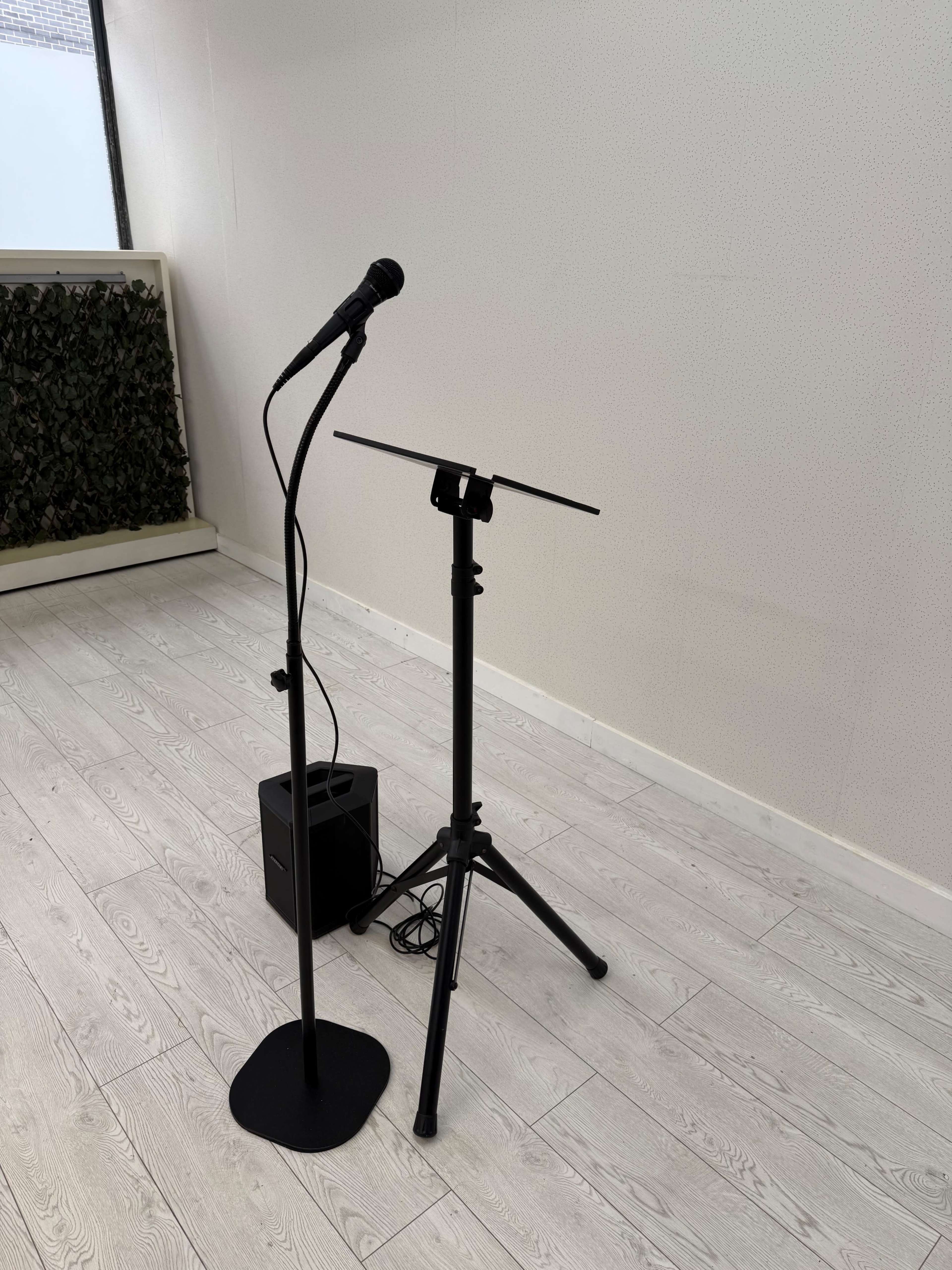 A black microphone on a stand is positioned next to a music stand in a minimalist room with light-colored wooden flooring.