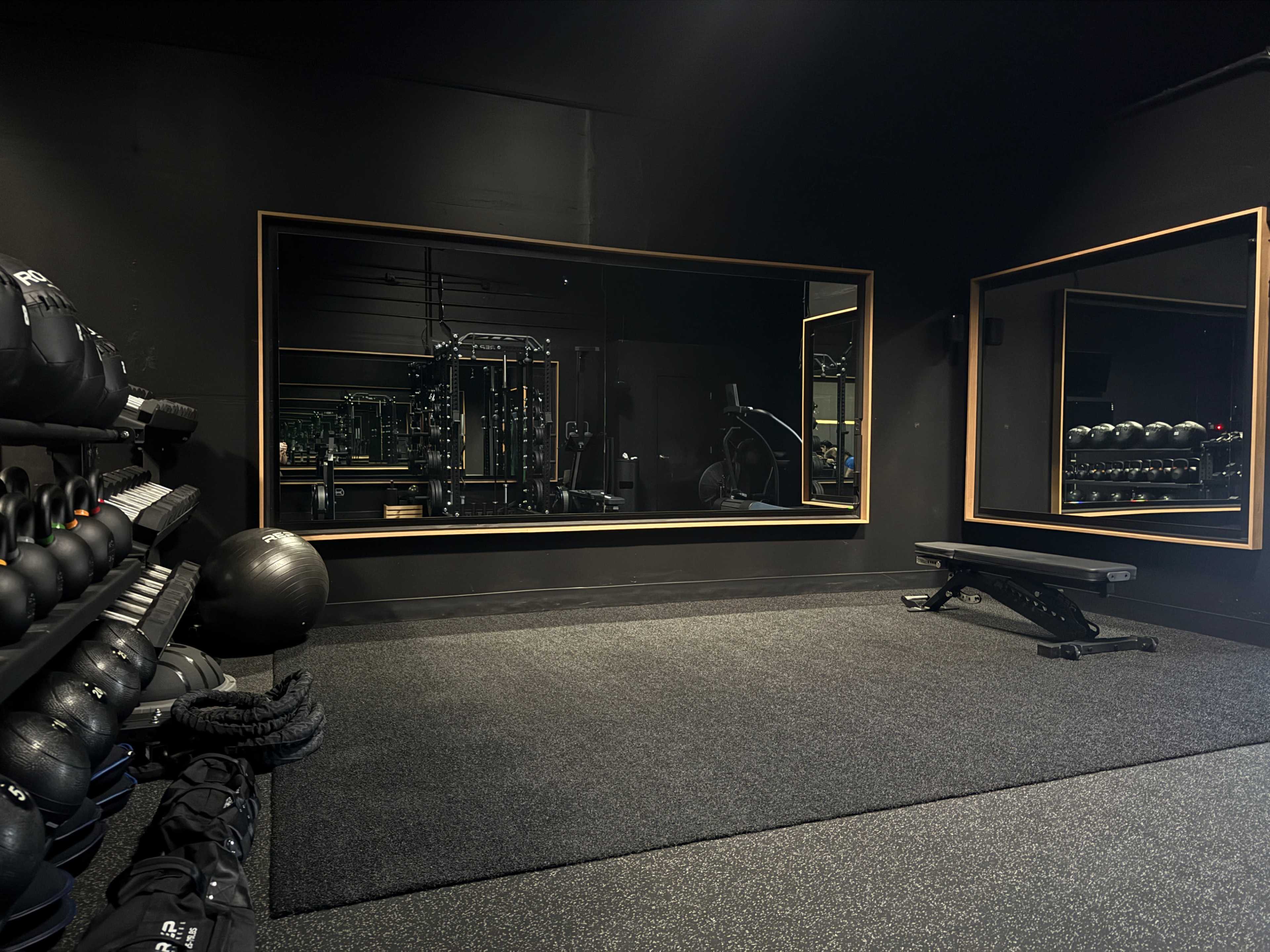 The image shows a small gym area with exercise equipment, a mirror, and a black rubber floor.