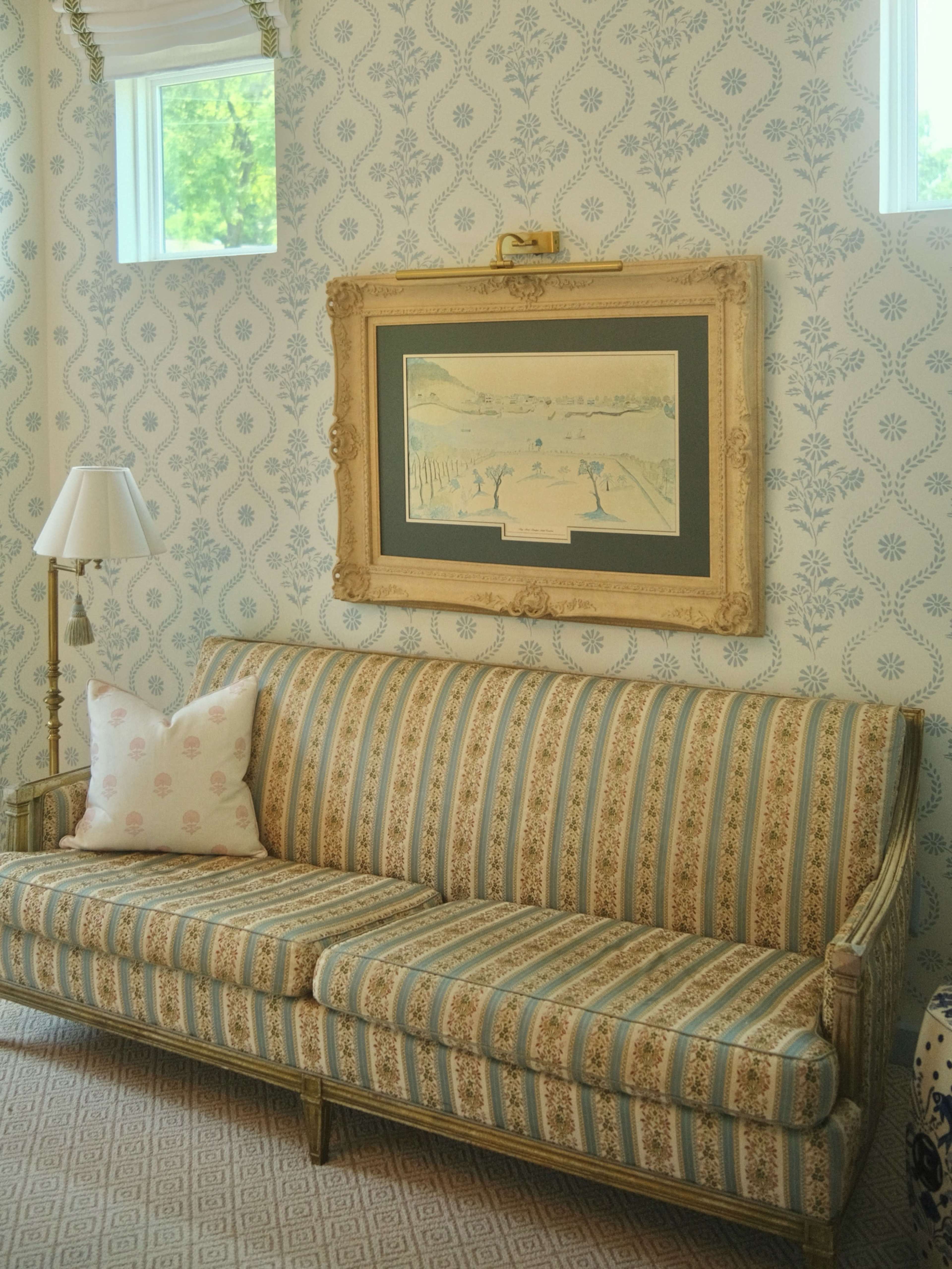 The image shows a patterned wallpapered room featuring a striped sofa with a decorative pillow, a framed artwork, and a lamp beside the couch.