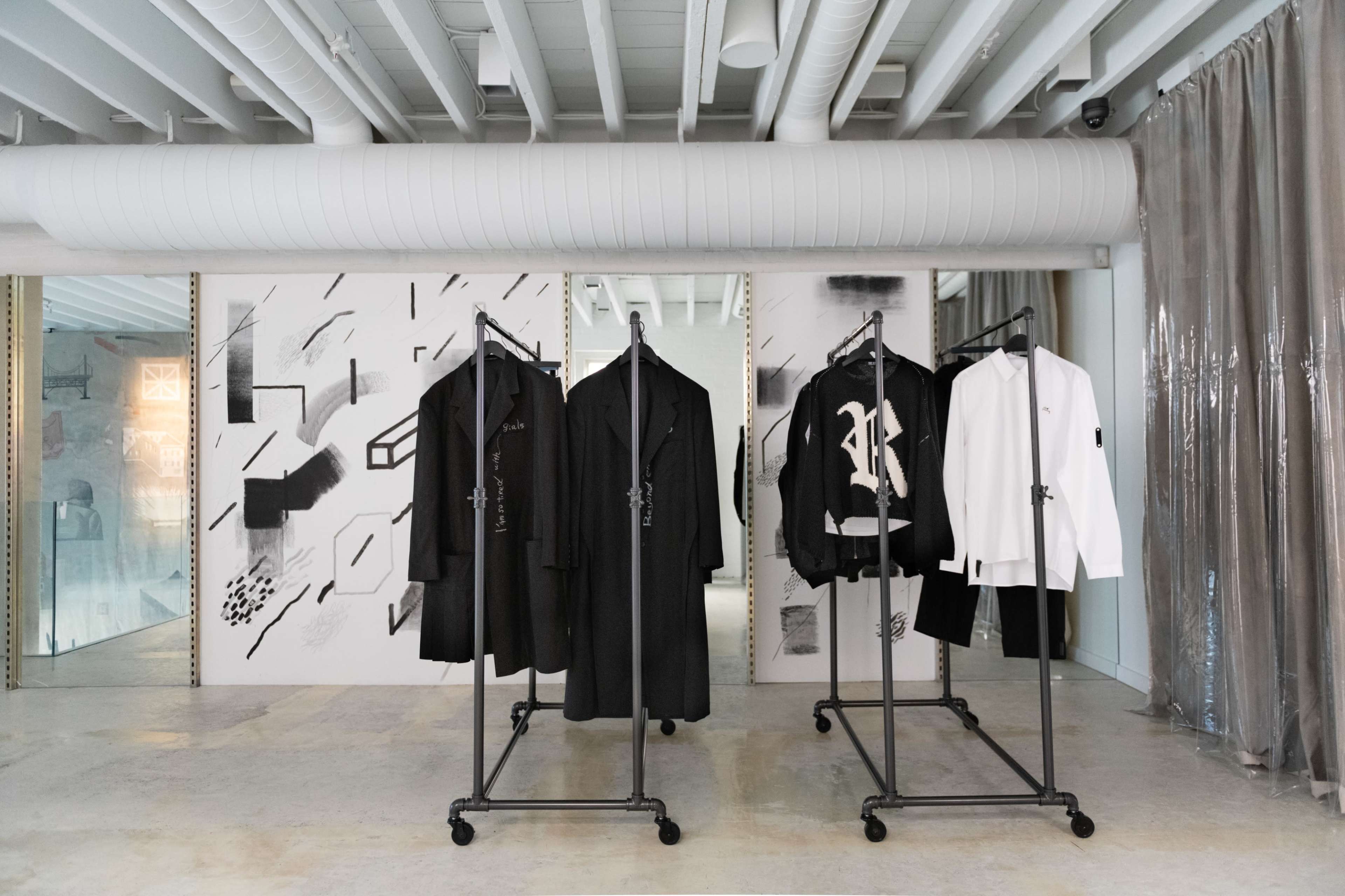 The image shows a modern clothing display featuring a series of black and white garments on rolling racks in a minimalist space with abstract wall art.