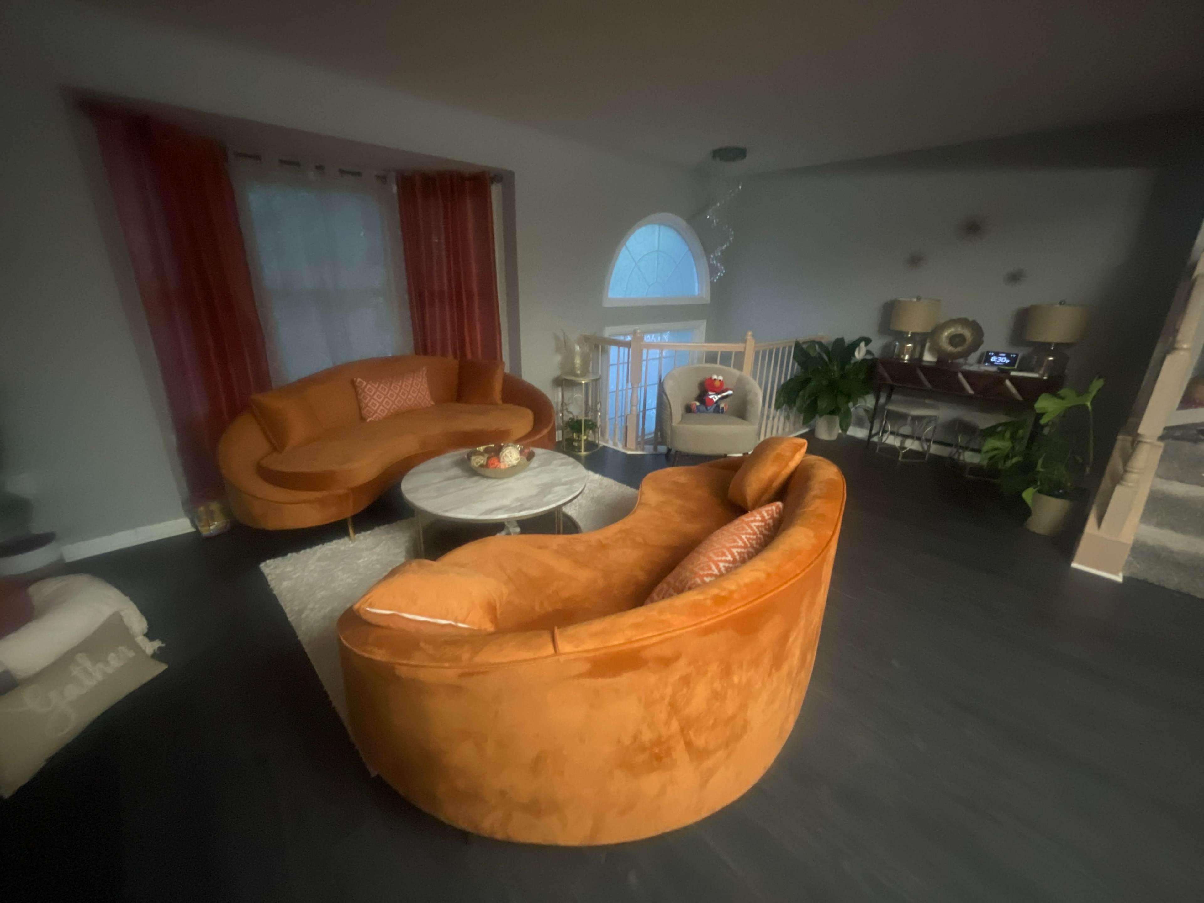 The image shows a living room featuring two orange sofas around a circular coffee table, with a light-colored rug and a staircase in the background.