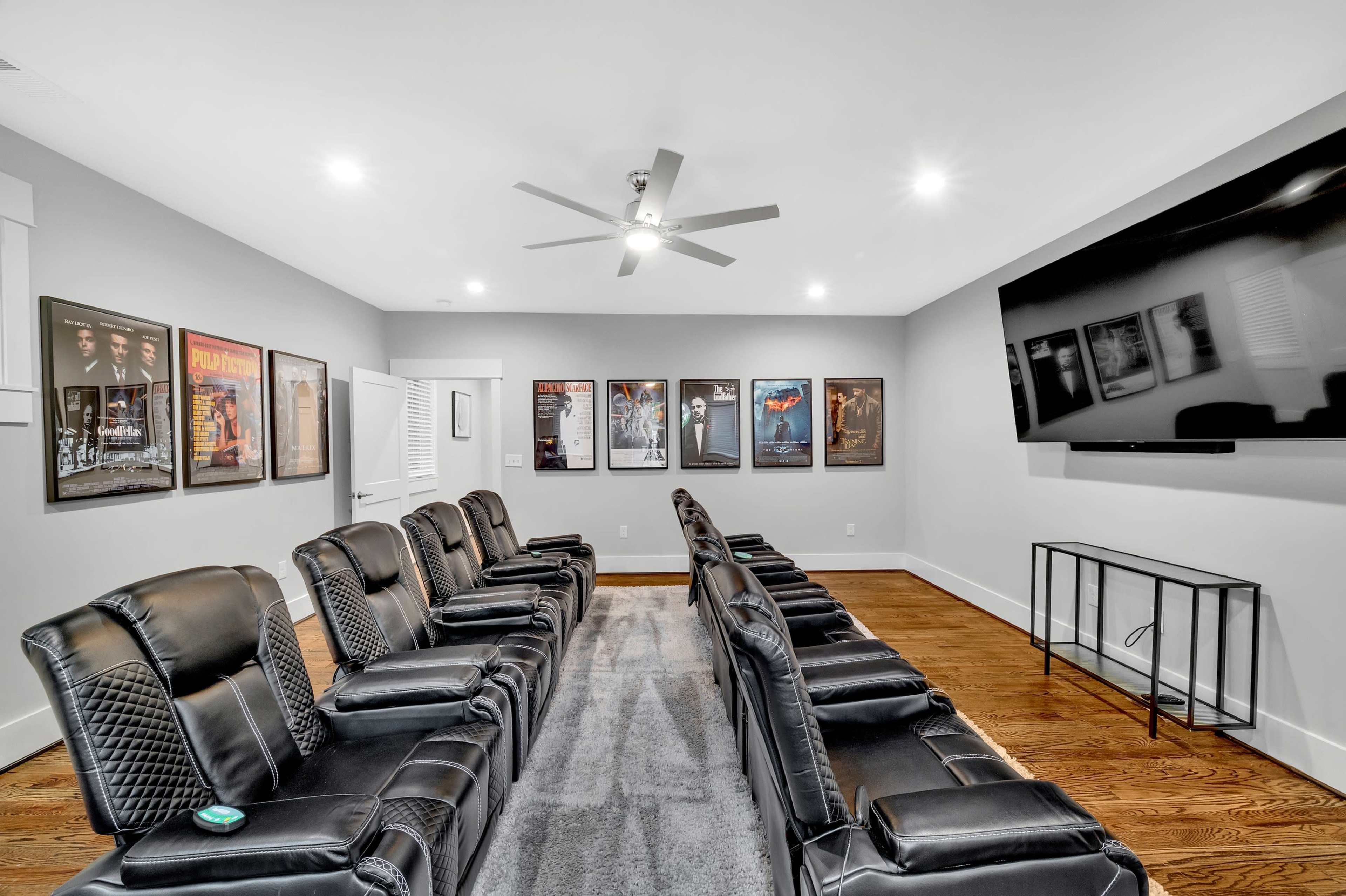 A home theater room features black reclining chairs arranged in rows, with movie posters on the walls and a large television mounted on one side.